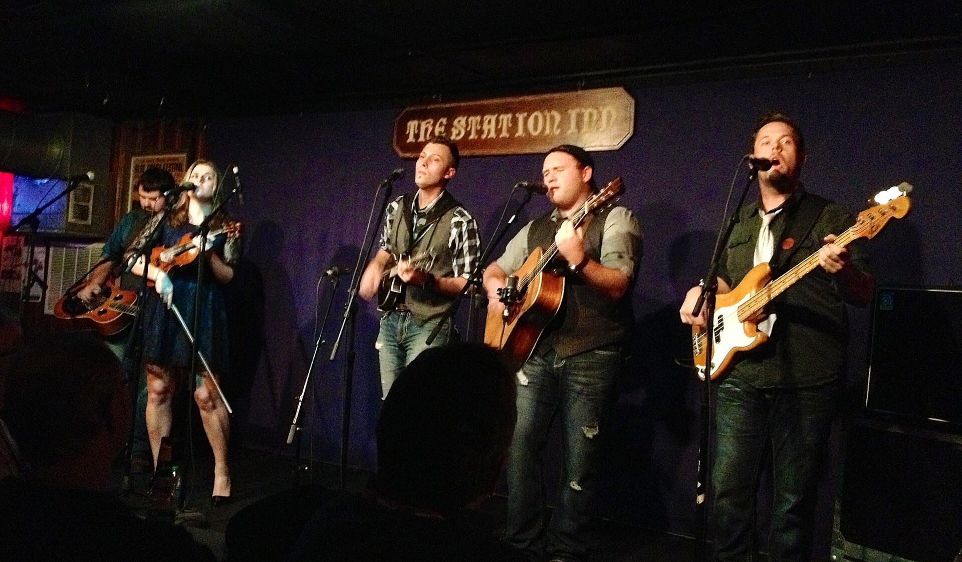 Cinco músicos actúan con guitarras, violín y bajo bajo el letrero de The Station Inn en un escenario tenuemente iluminado en Nashville.