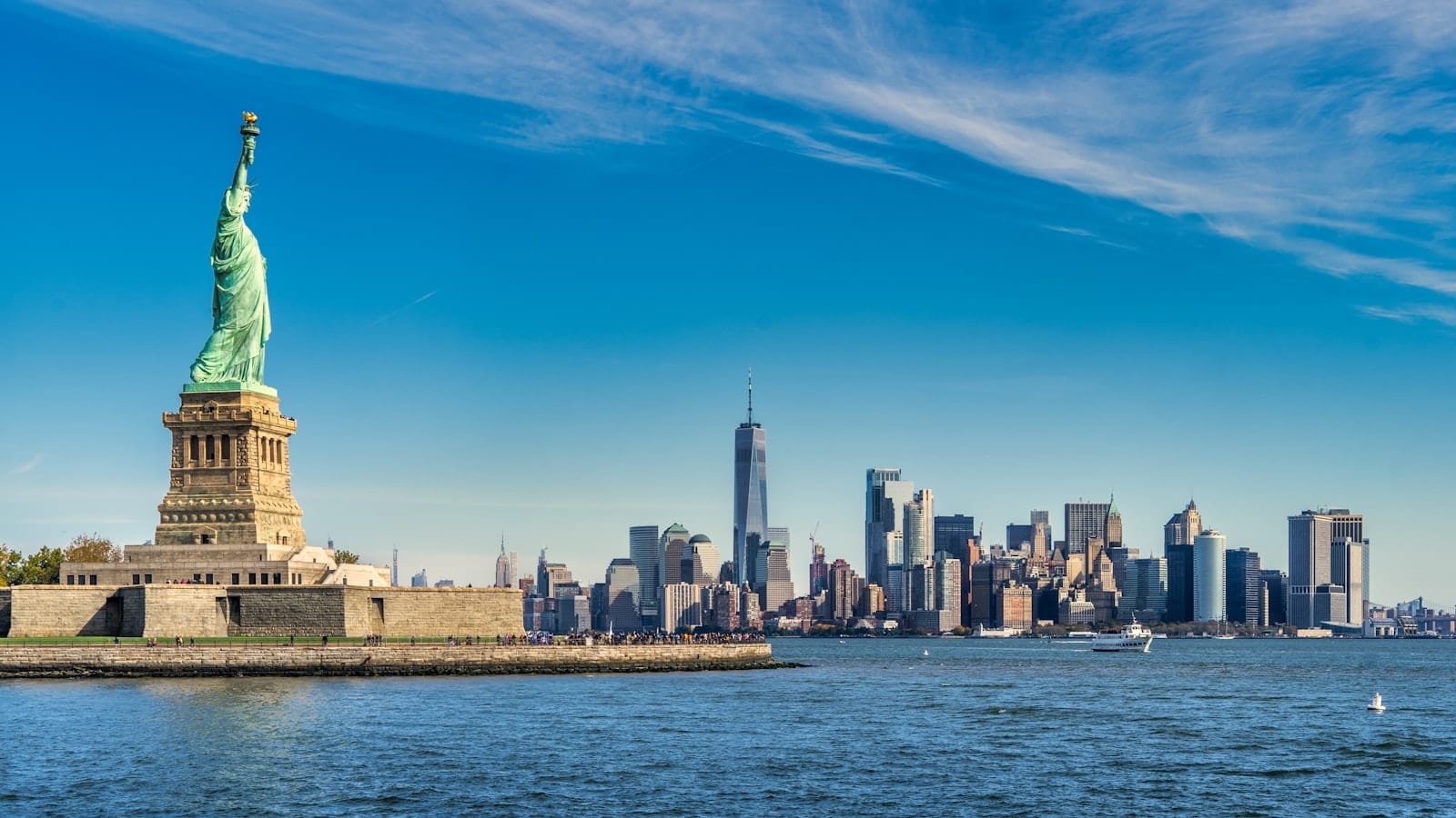La Statua della Libertà in primo piano con una vista nitida dello skyline di New York e il cielo azzurro sul porto.