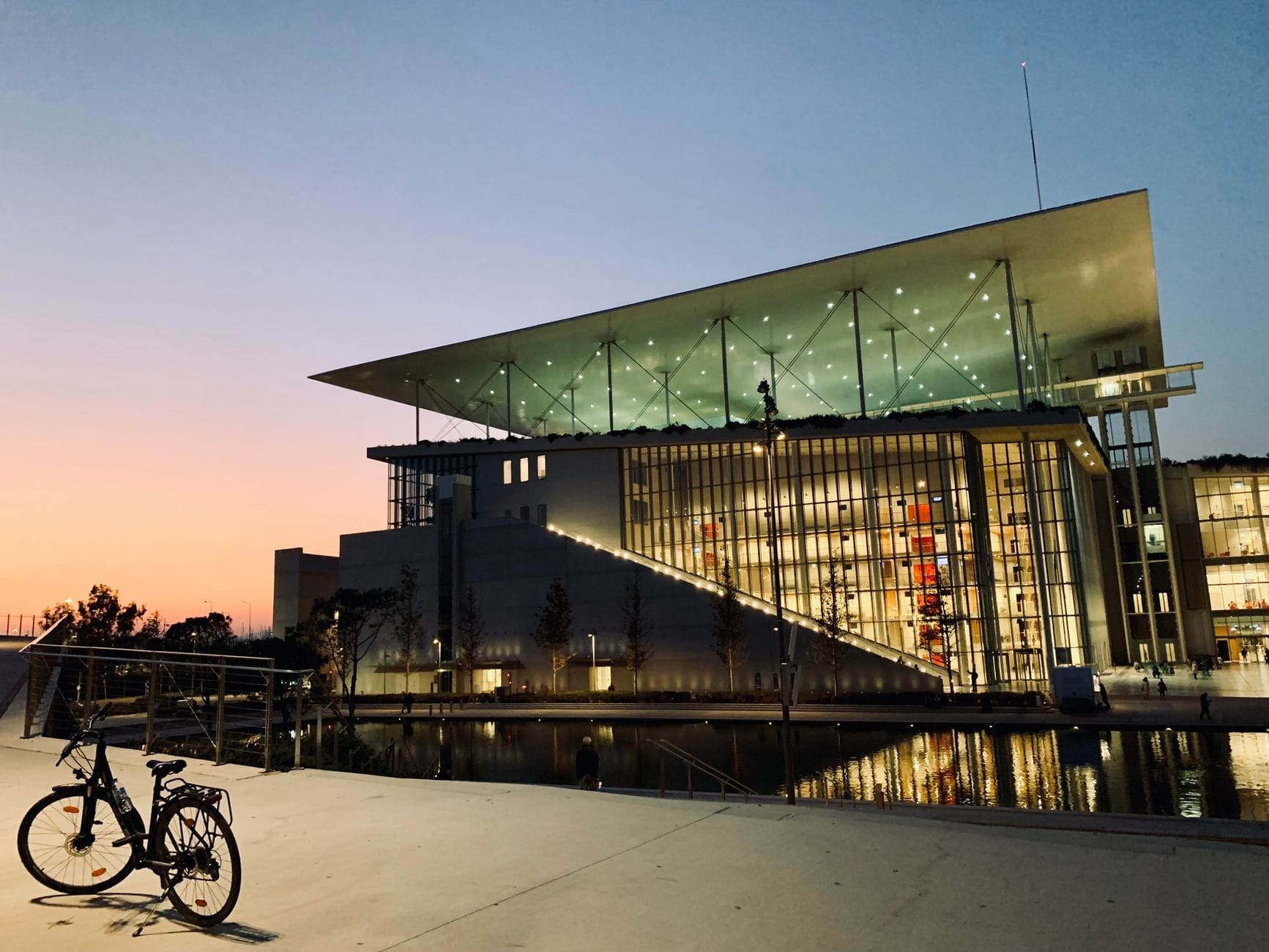 Vista serale dello Stavros Niarchos Foundation Cultural Center con la facciata in vetro illuminata, il caratteristico tetto inclinato e la vasca riflettente, sotto un cielo al tramonto variopinto ad Atene.