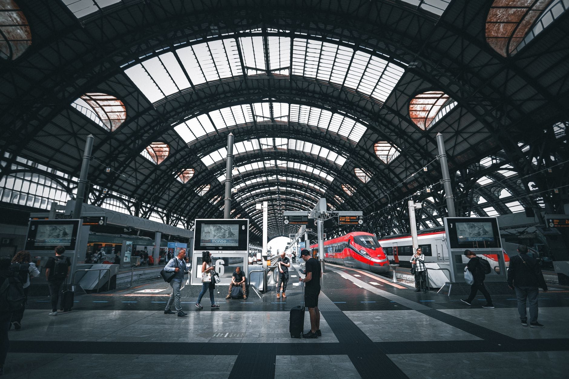 I viaggiatori camminano attraverso l'ampia piattaforma a volta in vetro e acciaio della Stazione di Milano Centrale, con un treno rosso ad alta velocità in attesa sotto il tetto ad arco.