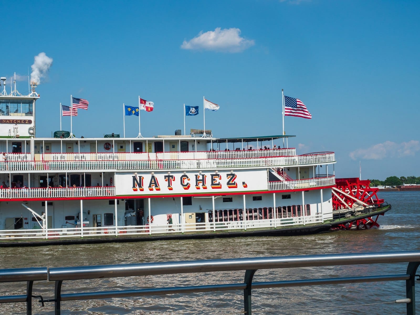 Il battello a vapore Natchez in navigazione sul Mississippi sotto un cielo azzurro limpido, con bandiere americane e passeggeri visibili su più ponti.