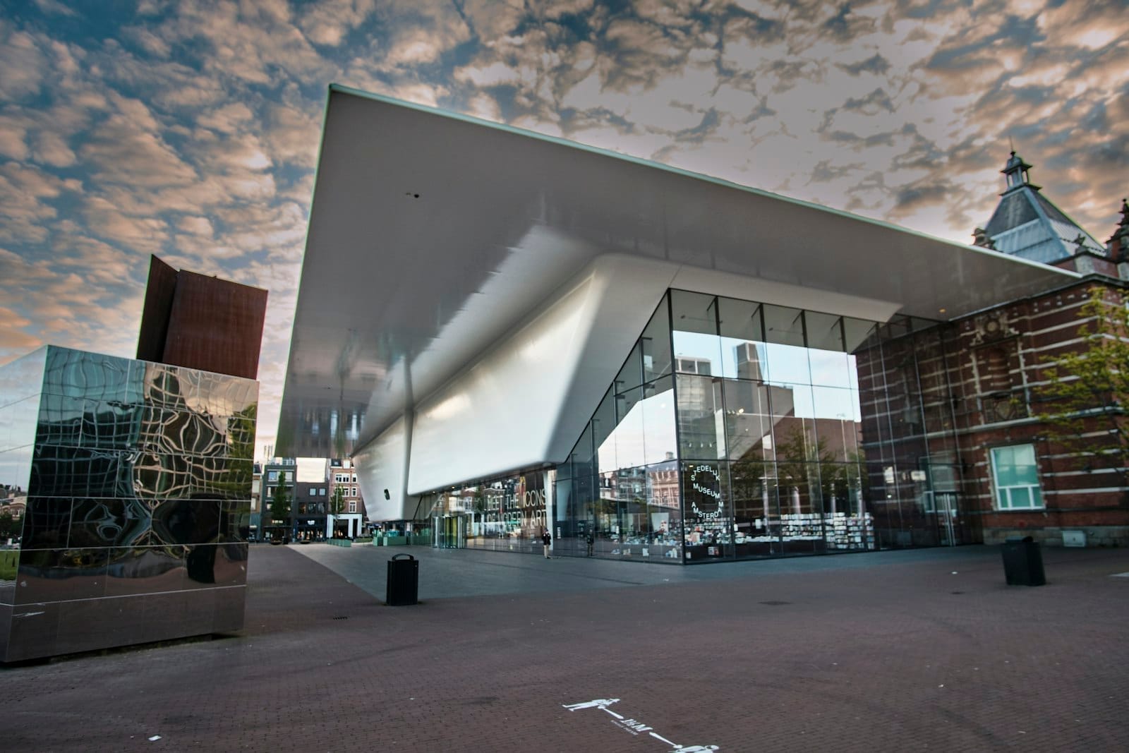Fachada moderna de cristal y blanca del Stedelijk Museum Amsterdam bajo un dramático cielo nublado, con reflejos en estructuras espejadas cercanas.