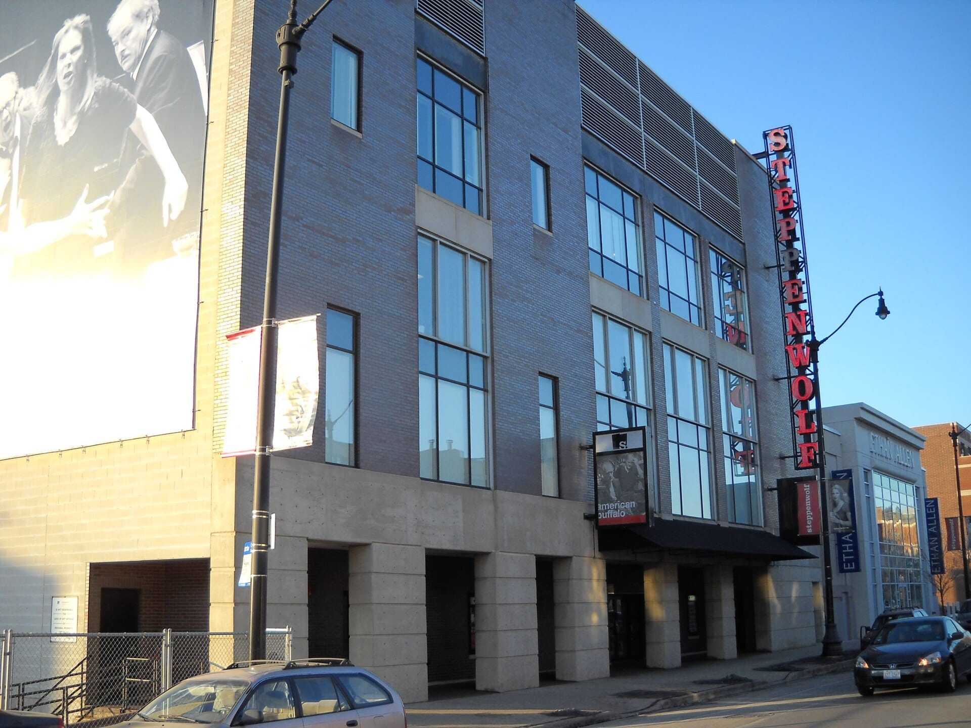 Vista esterna del moderno edificio dello Steppenwolf Theatre Company a Chicago, con grandi finestre, un'insegna rossa verticale e un murale in bianco e nero al tramonto.