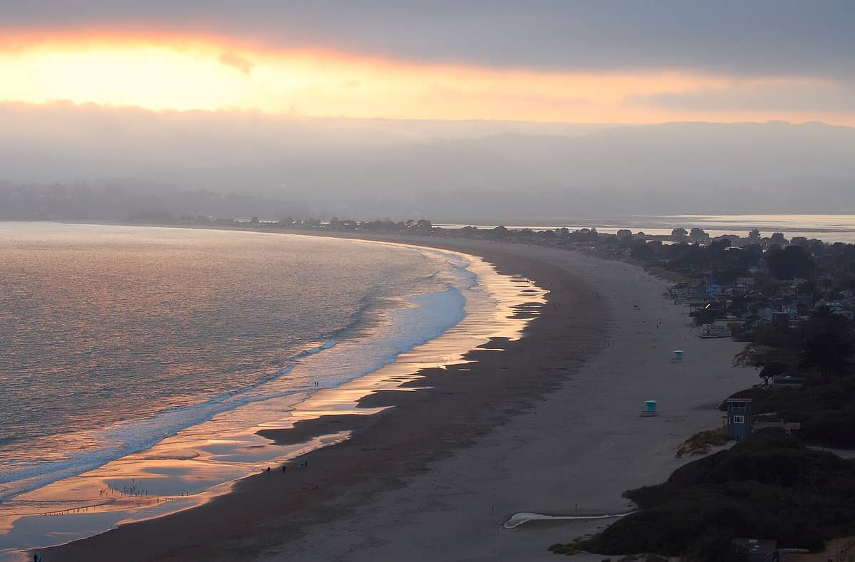 Ein breiter Sandstrand schwingt sich bei Sonnenuntergang entlang der Küste, mit sanften Wellen, weit entfernten Häusern und weichem Licht, das sich im Sand von Stinson Beach spiegelt.