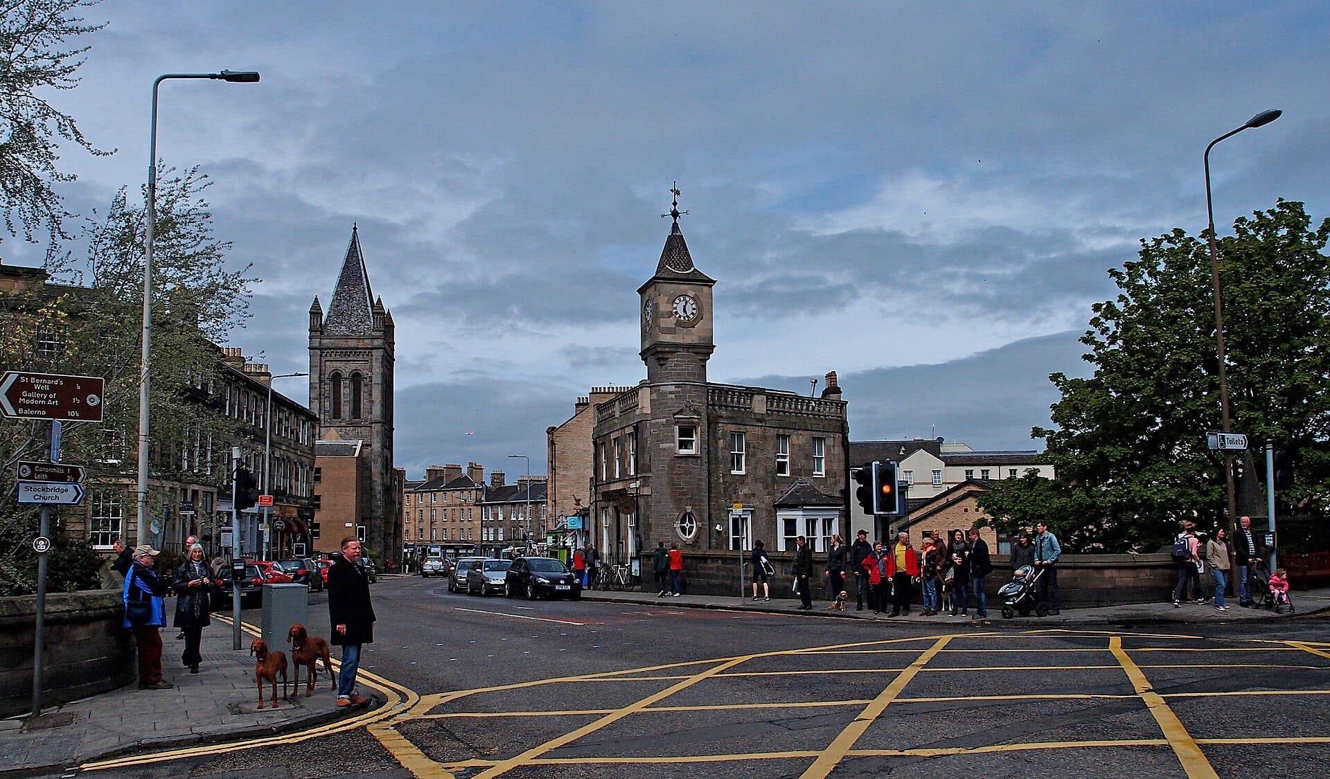 Kreuzung in Stockbridge, Edinburgh, mit historischen Steingebäuden, einem Kirchturm, einem Uhrenturm und Einheimischen, die an einem Fußgängerüberweg unter bewölktem Himmel warten.
