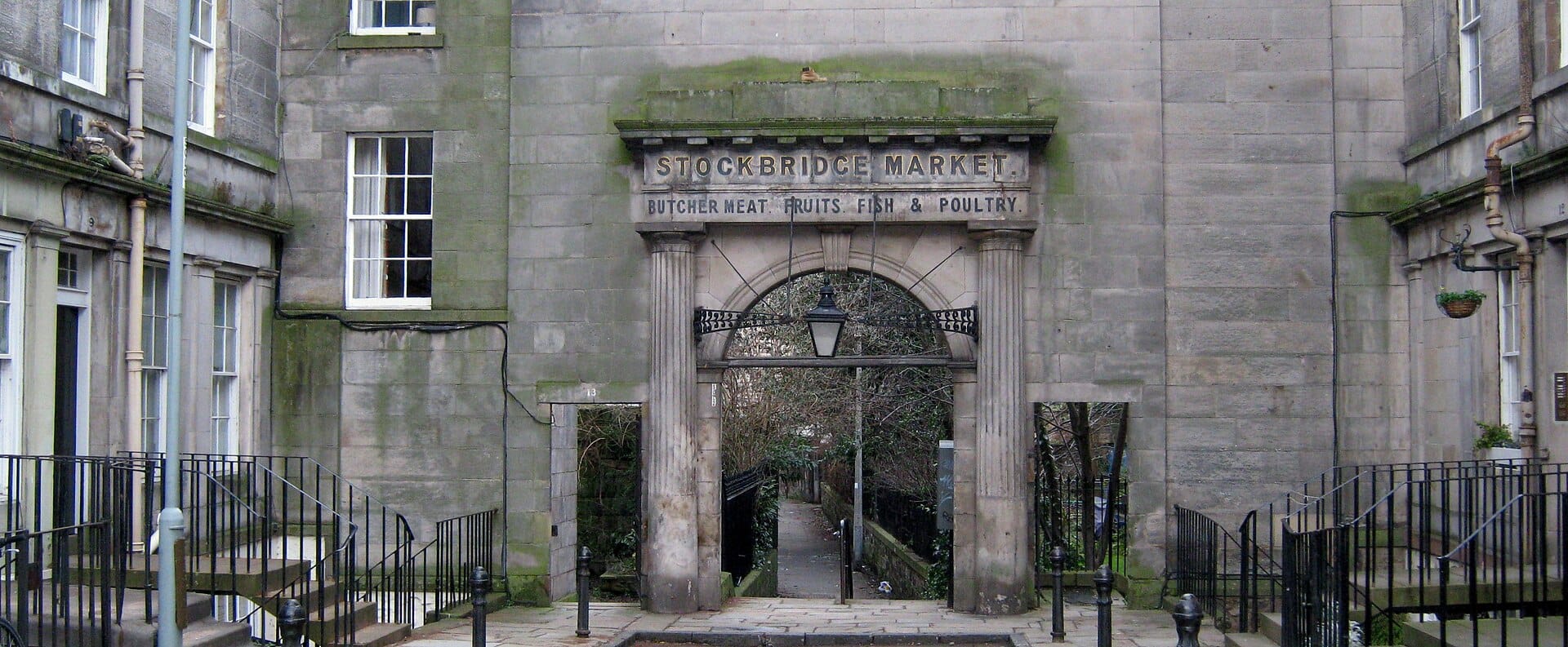 Das historische steinerne Eingangsportal des Stockbridge Market in Edinburgh, mit eingraviertem Marktnamen und Angebot sowie Wohngebäuden auf beiden Seiten.