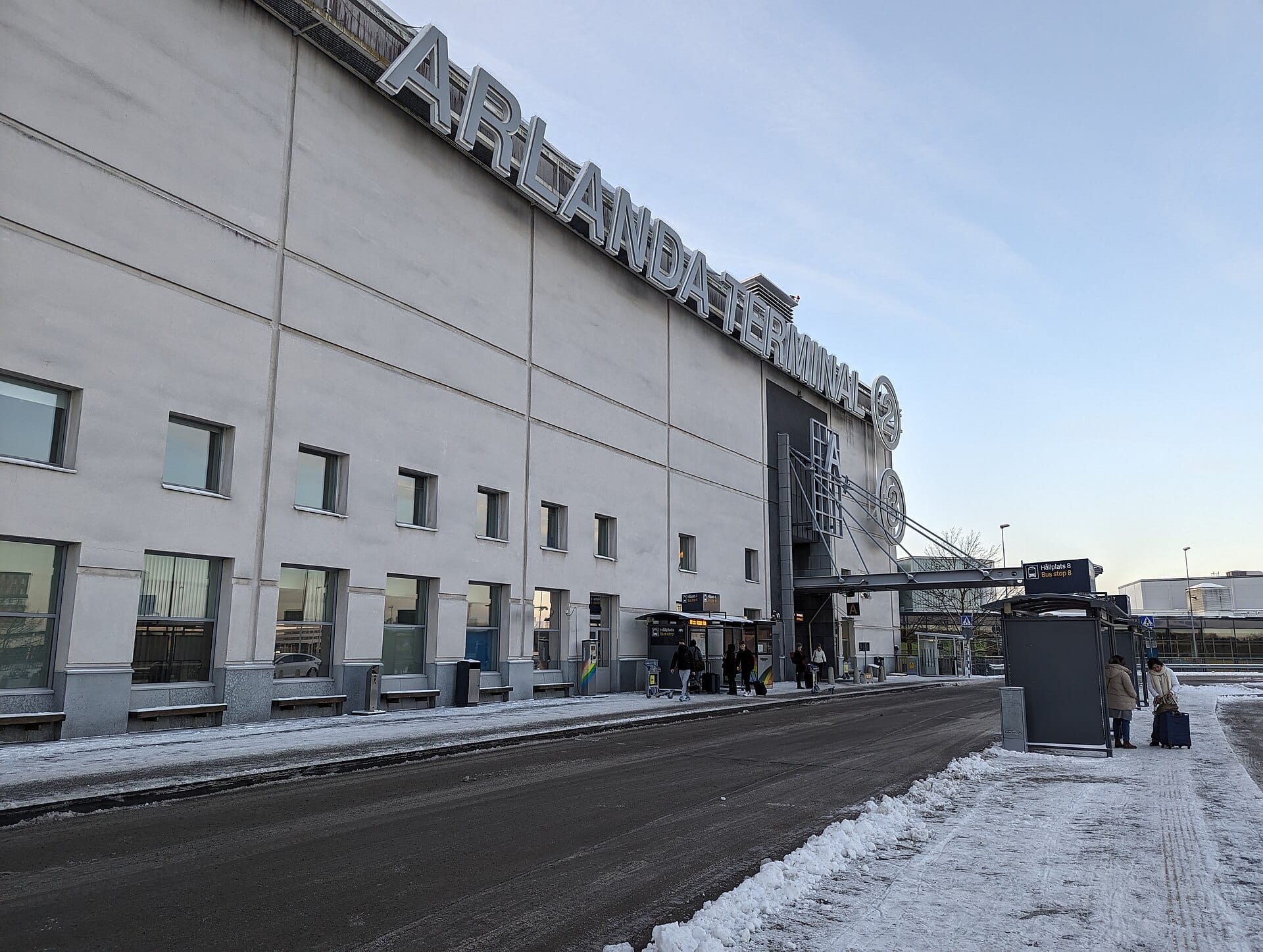วิวภายนอกของอาคารผู้โดยสาร 4 สนามบิน Stockholm Arlanda พร้อมป้ายอาคารขนาดใหญ่ พื้นปกคลุมด้วยหิมะ ป้ายรถประจำทาง และนักเดินทางด้านนอก