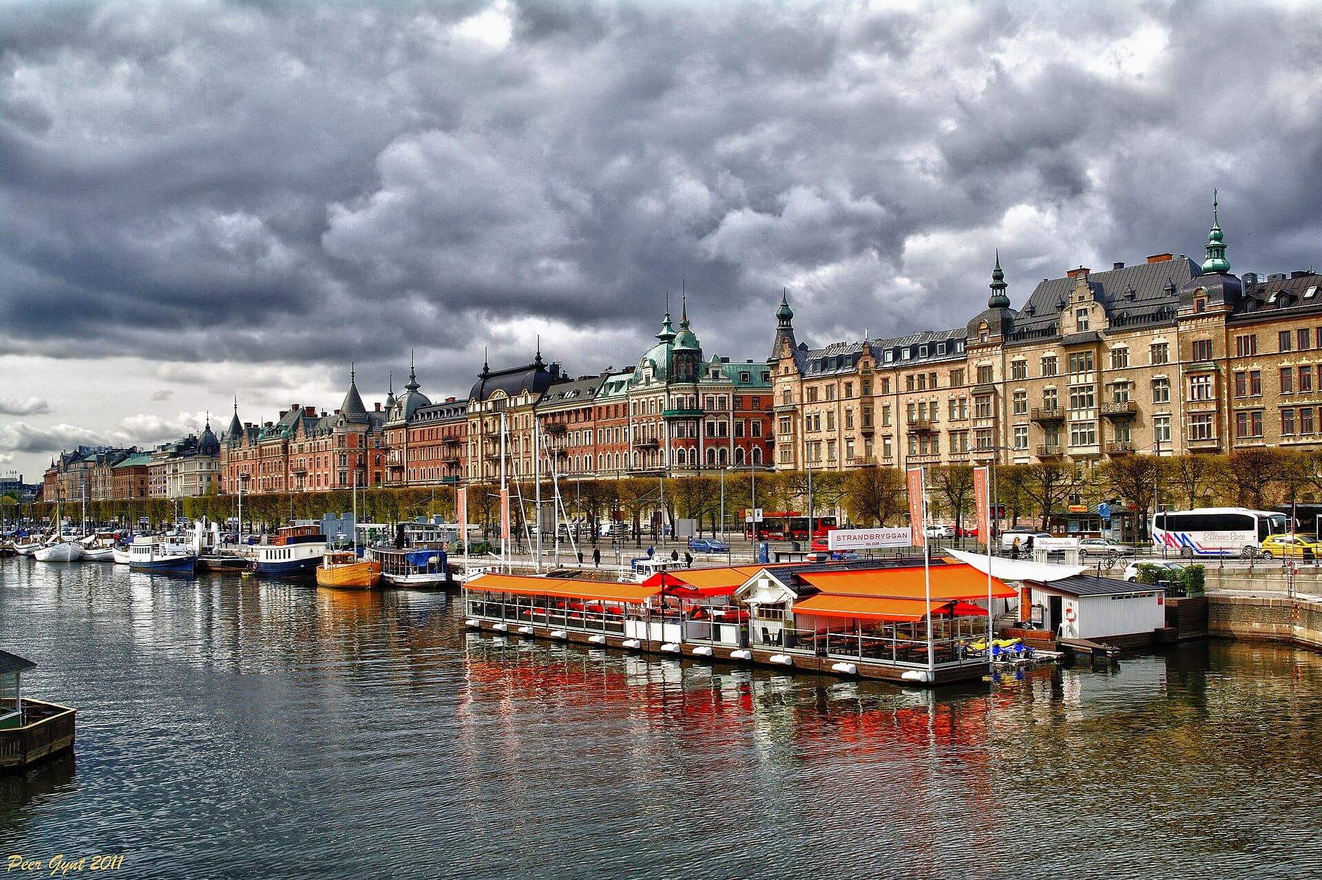 อาคารประวัติศาสตร์หลากสีสันตามแนวริมน้ำ Strandvägen ในย่าน Östermalm ของสตอกโฮล์ม พร้อมเรือและร้านอาหารลอยน้ำใต้ท้องฟ้าที่มีเมฆดราม่า