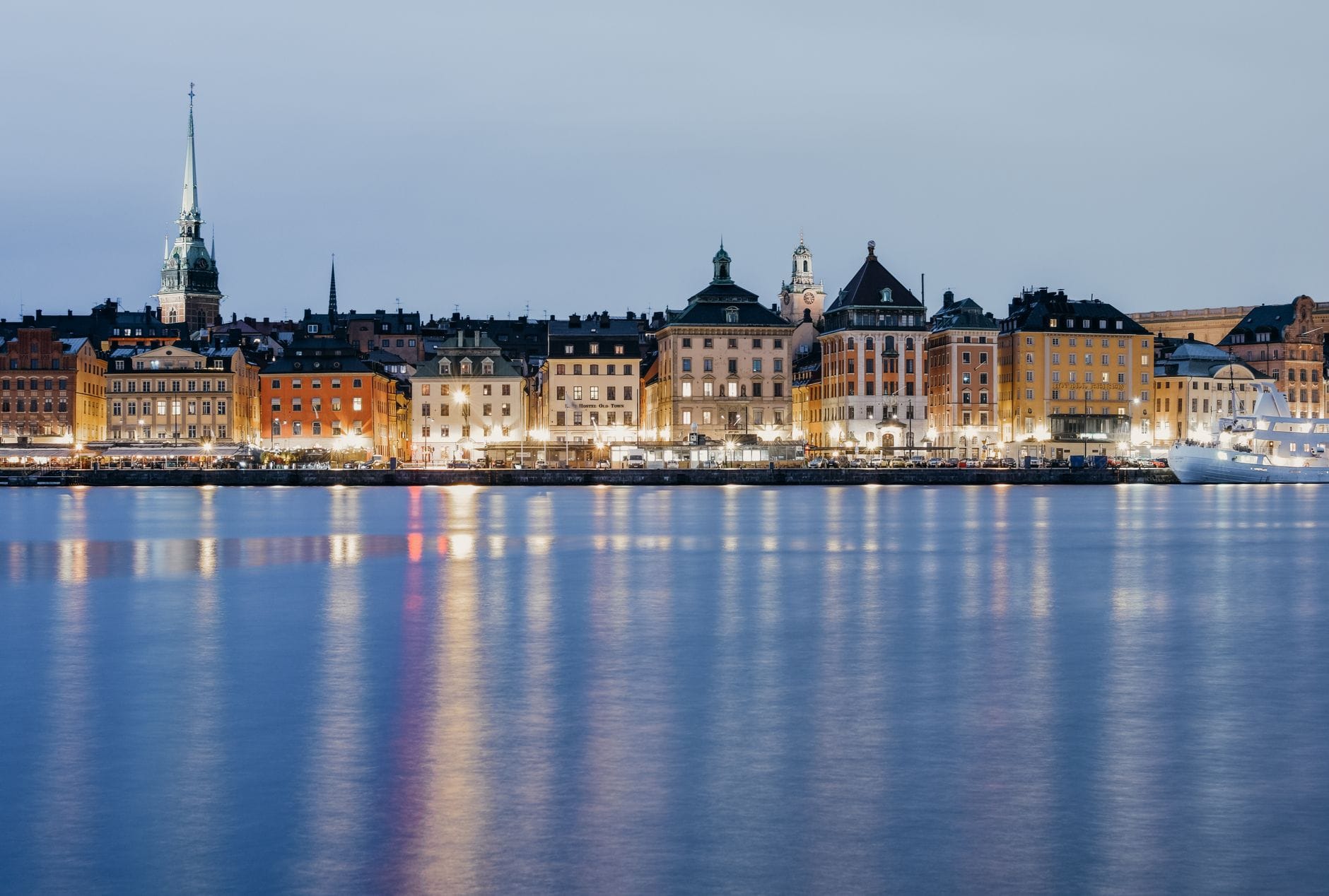 วิวพาโนรามาตอนเย็นของ Gamla Stan ริมน้ำในสตอกโฮล์ม ตึกราบ้านช่องโบราณและยอดโบสถ์สะท้อนเงาในผืนน้ำสงบ ท้องฟ้าแจ่มใส