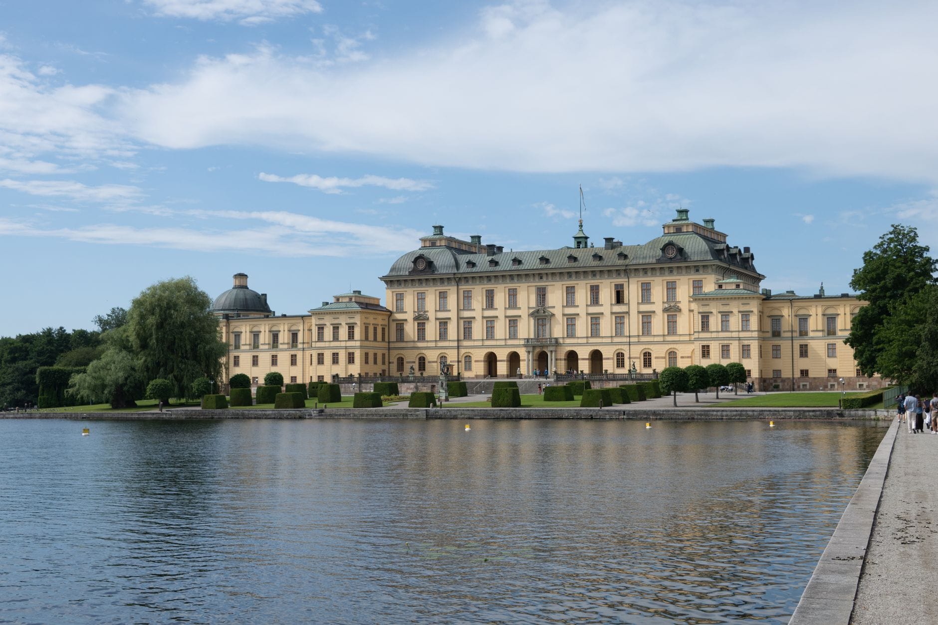 วิวทิวทัศน์ของพระราชวัง Drottningholm ริมน้ำในวันฤดูร้อนที่แดดส่อง พร้อมสวนและท้องฟ้าสีฟ้าใส
