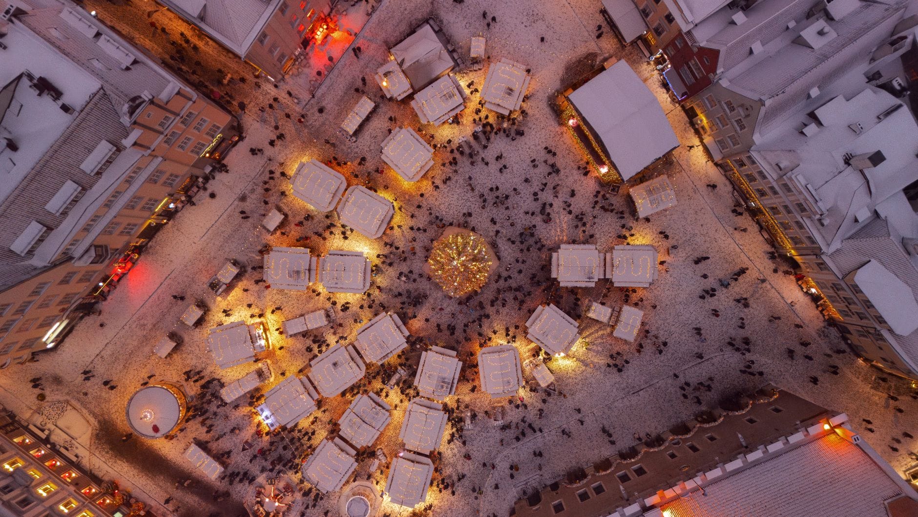 ภาพมุมสูงของจัตุรัส Stortorget ในสตอกโฮล์มยามพลบค่ำ เต็มไปด้วยแผงตลาดคริสต์มาส ฝูงชน และแสงไฟเทศกาล ล้อมรอบด้วยอาคารประวัติศาสตร์ที่ปกคลุมด้วยหิมะเล็กน้อย