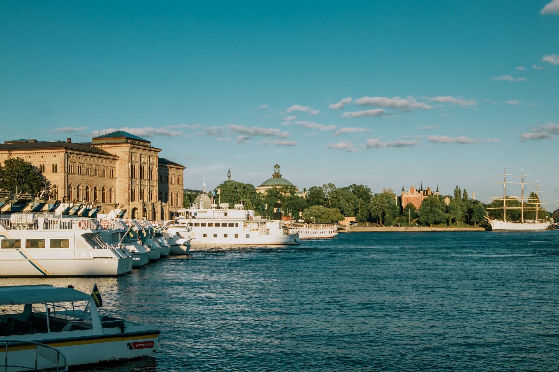 ริมน้ำของ Stockholm ในยามเย็นฤดูร้อน พร้อมเรือจอดเทียบท่า อาคารประวัติศาสตร์ ต้นไม้เขียวขจี และท้องฟ้าสีฟ้าใส สื่อถึงบรรยากาศตามฤดูกาลที่มีชีวิตชีวา