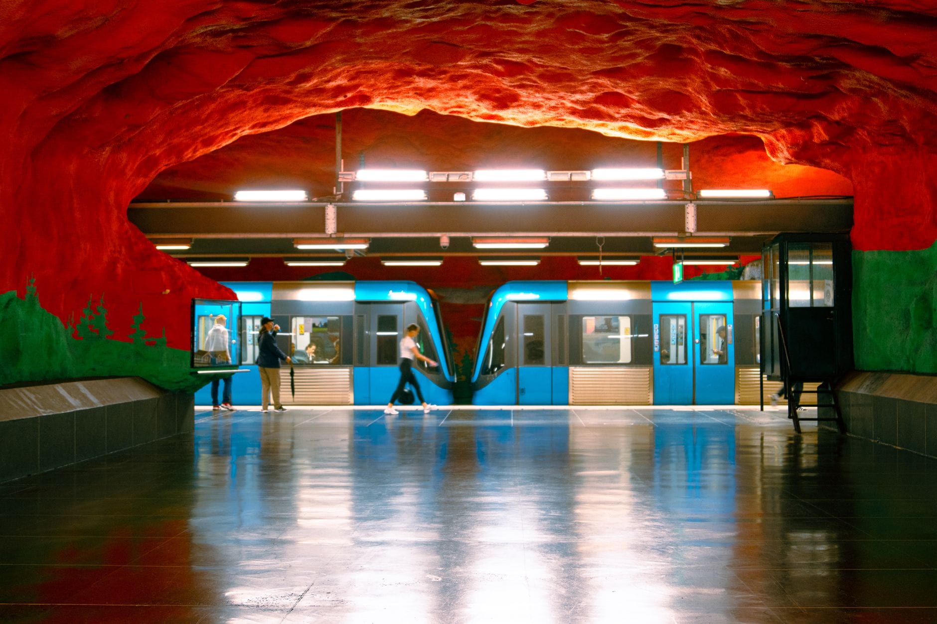 สถานีรถไฟใต้ดินสตอกโฮล์มที่มีเพดานสีแดงรูปทรงถ้ำอันเป็นเอกลักษณ์ รถไฟใต้ดินสีน้ำเงิน และผู้คนบนชานชาลา
