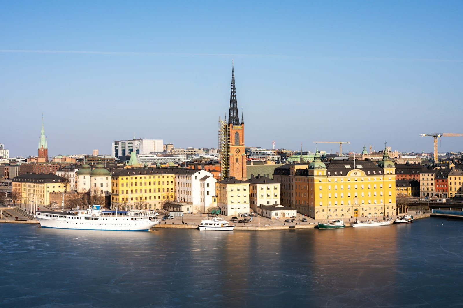 Panoramic view of Stockholm’s Gamla Stan waterfront, featuring colorful historic buildings and the Riddarholmen Church with its distinctive spire, under a clear blue sky.