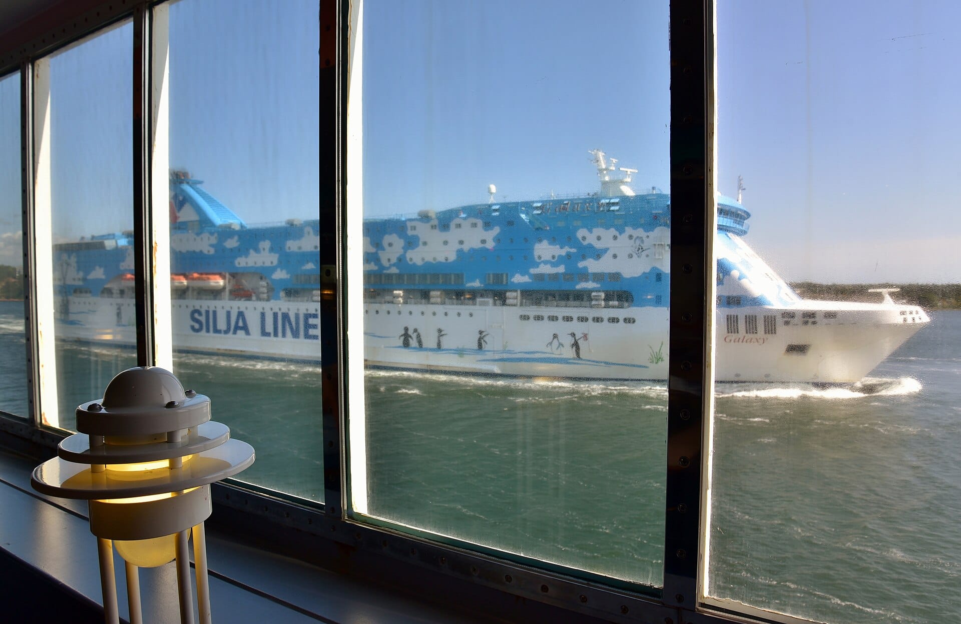 วิวจากหน้าต่างของเรือเฟอร์รี่ แสดงให้เห็นเรือสำราญขนาดใหญ่ Silja Line Galaxy แล่นผ่านไปบนน้ำภายใต้ท้องฟ้าสีฟ้าใส