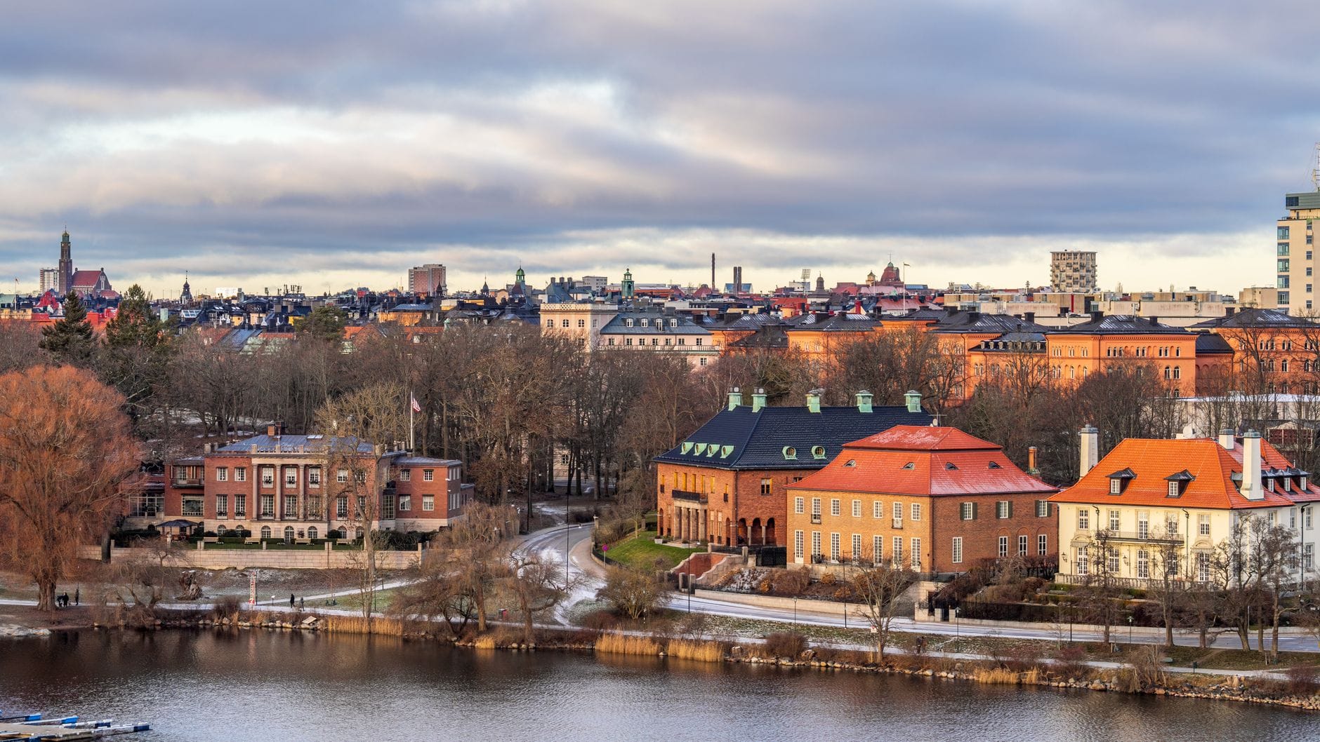วิวเกาะ Djurgården พร้อมอาคารประวัติศาสตร์ริมน้ำ ต้นไม้ และเส้นขอบฟ้าของ Stockholm ในฉากหลังในวันที่มีเมฆมาก