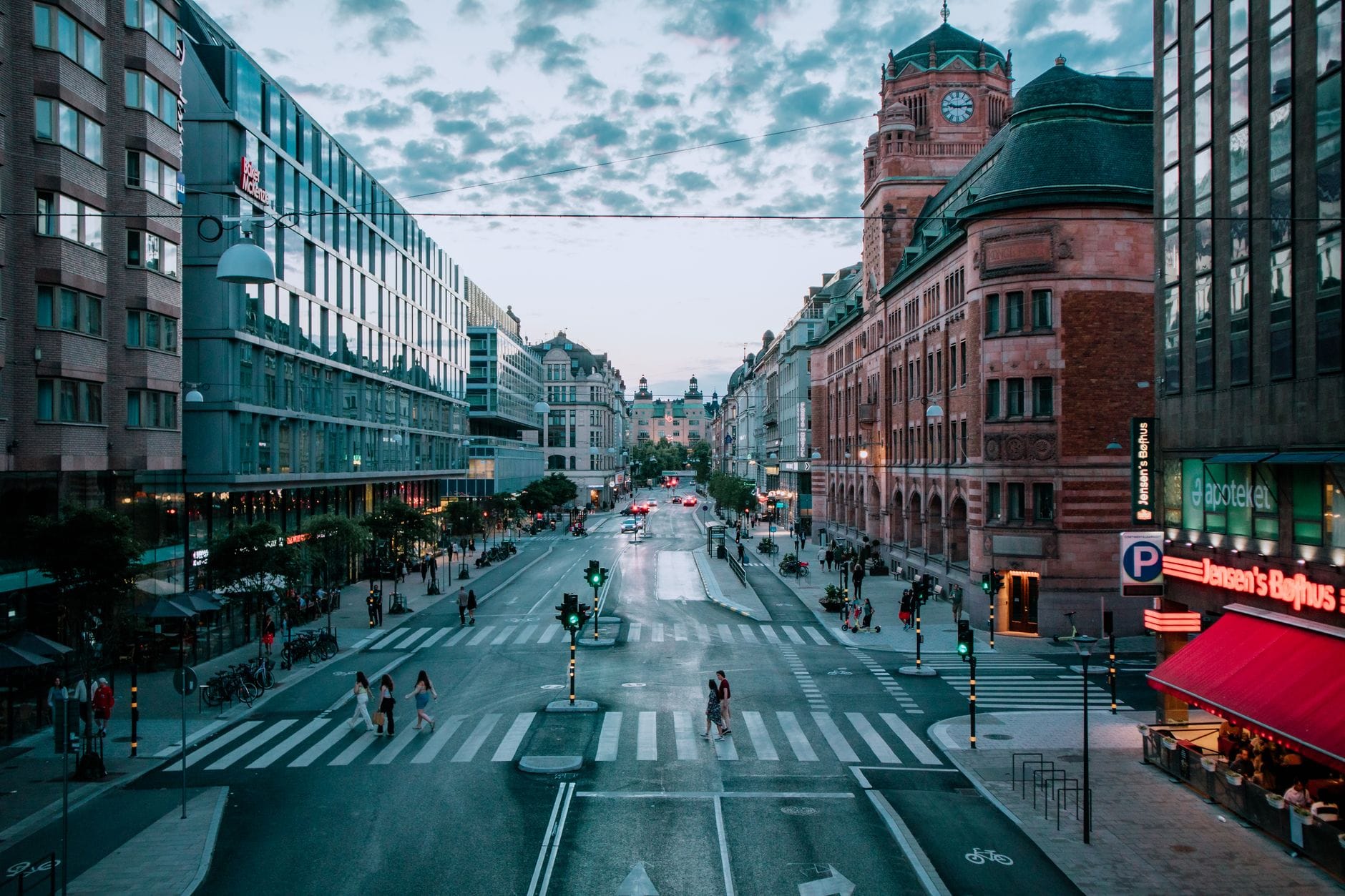 วิวกว้างของถนนสายหลักในใจกลาง Stockholm ที่คึกคักพร้อมทางม้าลาย ผู้คนเดินผ่าน และอาคารประวัติศาสตร์ทั้งสองข้างในยามพลบค่ำ