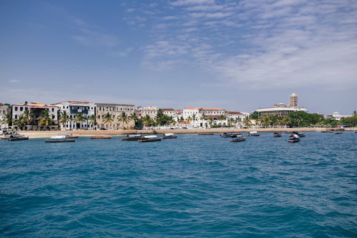 Weitwinkelansicht der Uferpromenade von Stone Town in Sansibar mit vertäuten Booten, Kolonialgebäuden und blauem Himmel, tagsüber vom Meer aus gesehen.