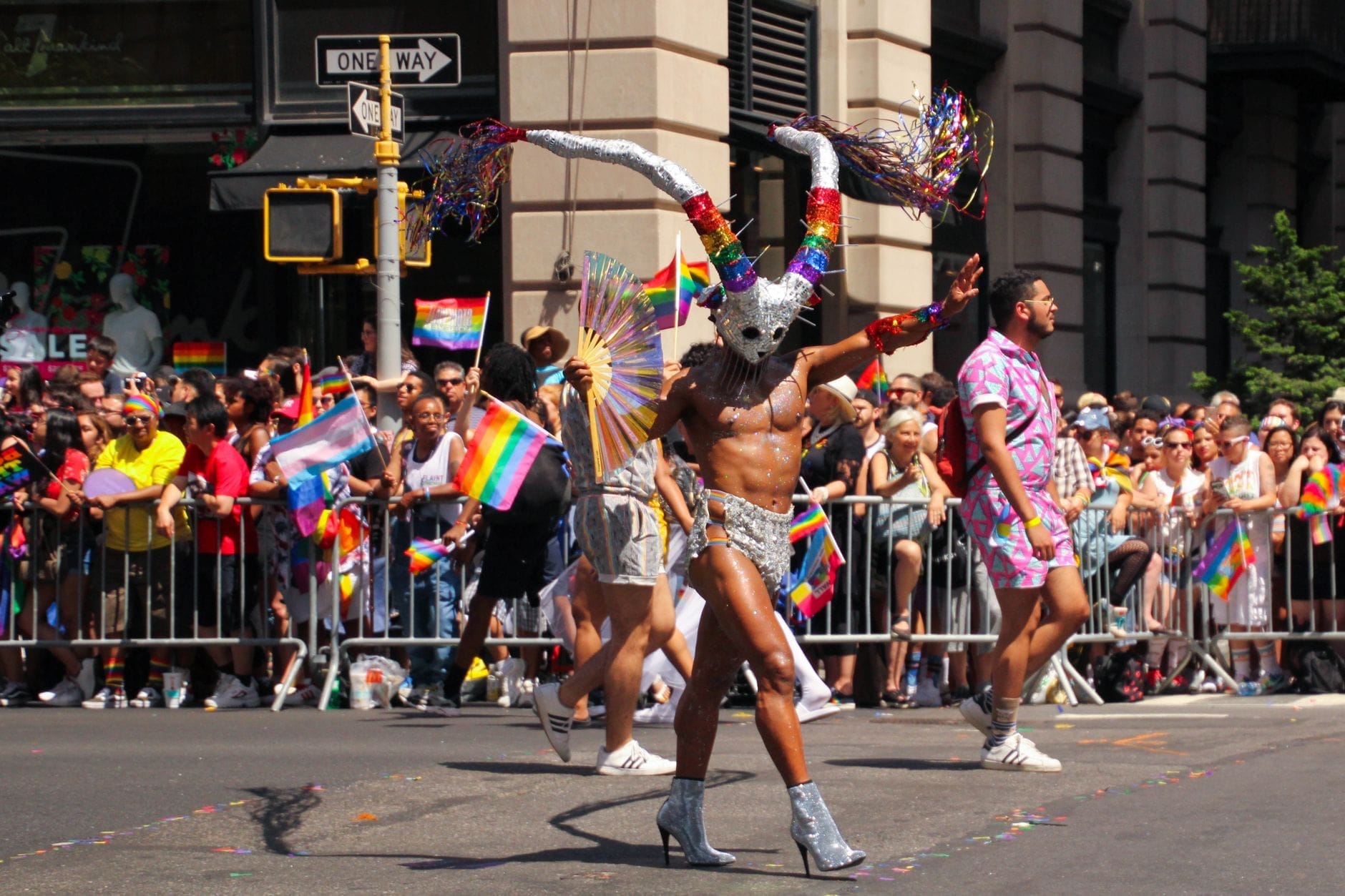 Colorata parata del Pride con un ballerino in costume e bandiere arcobaleno, folle radunate per strada a celebrare la cultura e la comunità LGBTQ di giorno.
