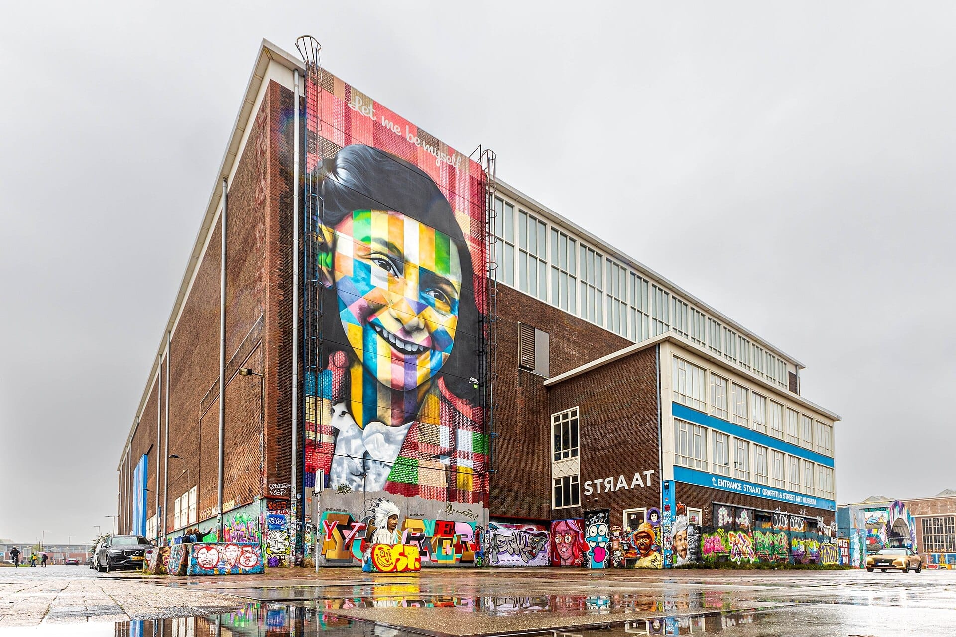 Gran almacén de ladrillo del STRAAT Museum en Ámsterdam, con murales vibrantes y un colorido retrato en un día lluvioso y nublado.