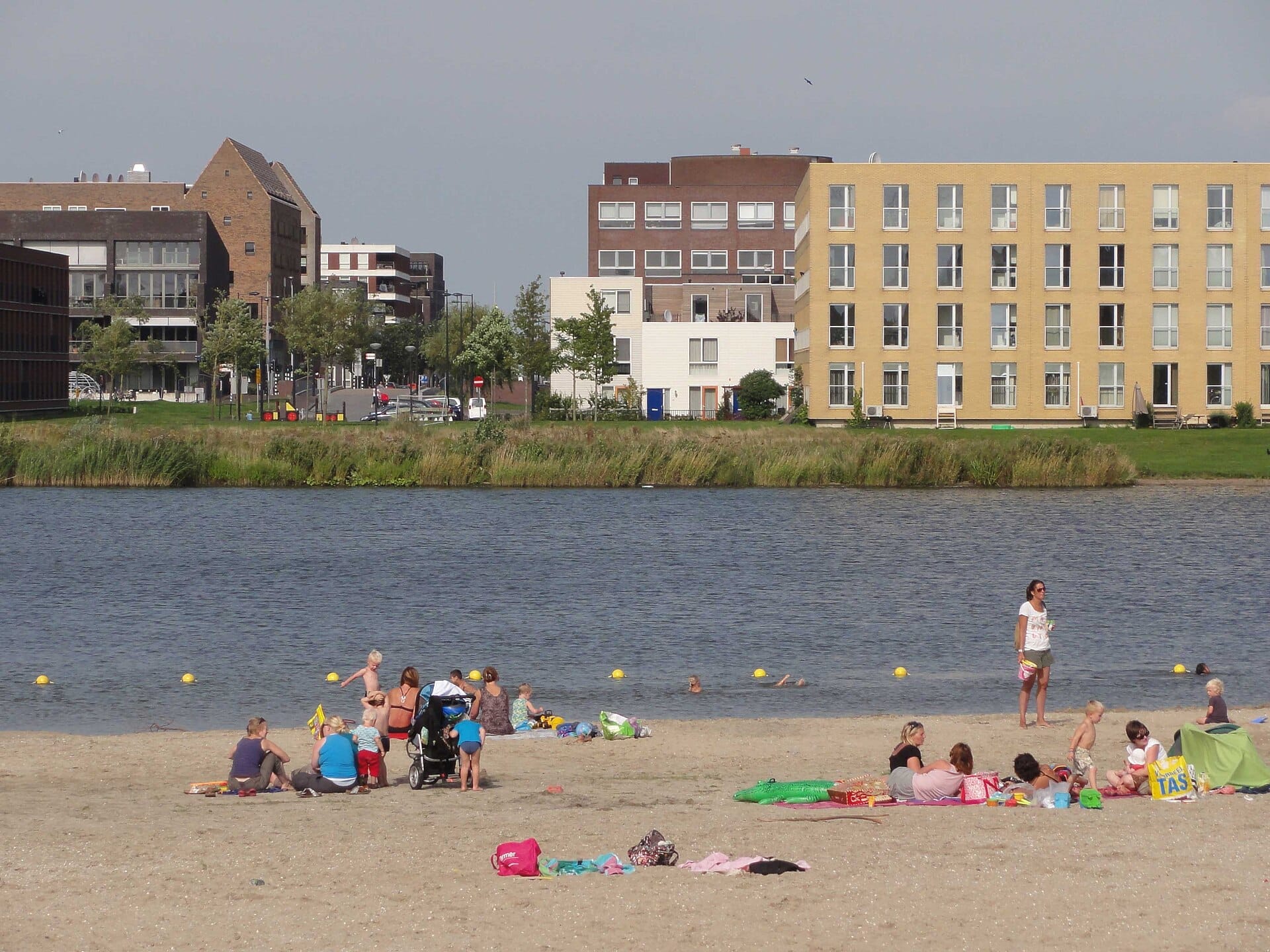 Familias y niños se relajan en la orilla arenosa de Strand IJburg, con modernos edificios de apartamentos y aguas tranquilas de fondo.