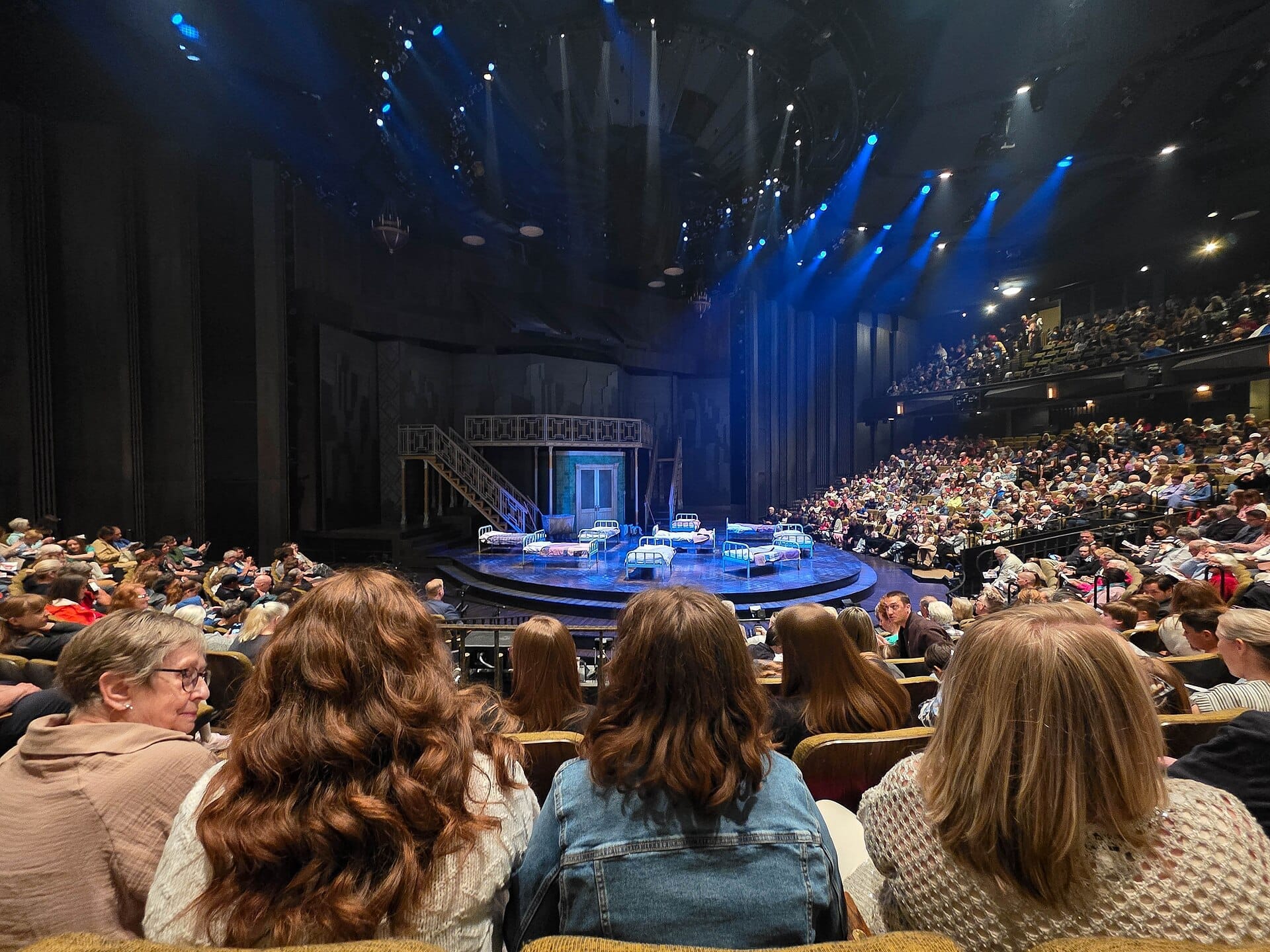 ผู้ชมเต็มโรงละคร Stratford Festival หันหน้าสู่เวทีทรงกลมที่ตกแต่งด้วยเตียงและแสงไฟสีน้ำเงินก่อนเริ่มการแสดง