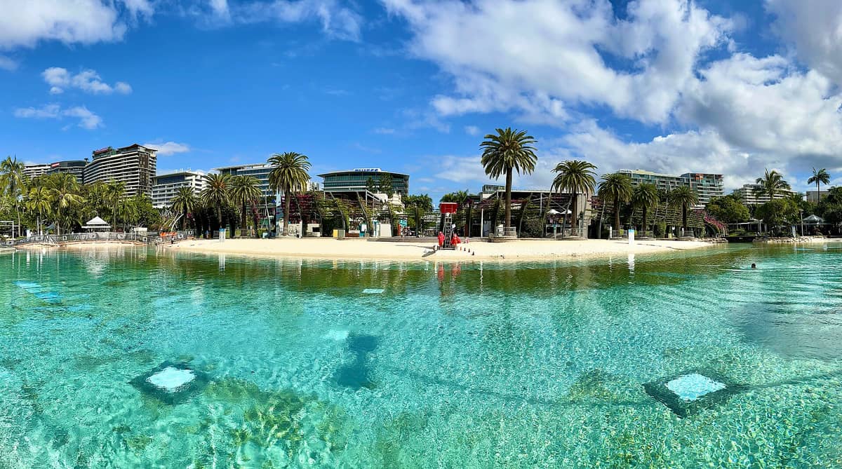 Wide landscape view of Streets Beach at South Bank Parklands in Brisbane, featuring turquoise lagoon water, sandy beach, palm trees, lifeguard, and city buildings in background under a blue sky.