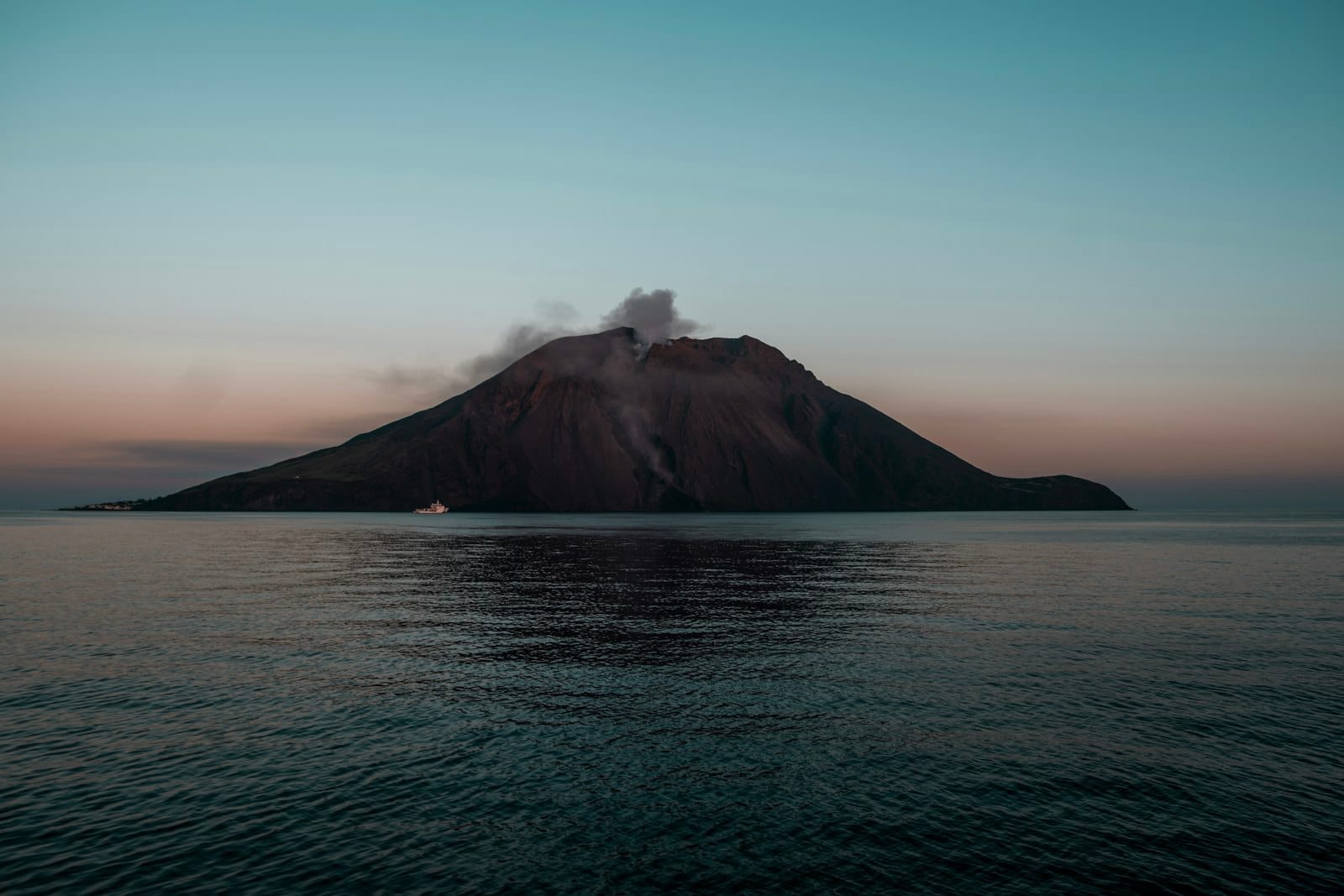 Vulkan Stromboli mit aufsteigendem Rauch aus seinem Krater, bei Abenddämmerung über ruhigem Wasser mit einem Boot in Ufernähe.