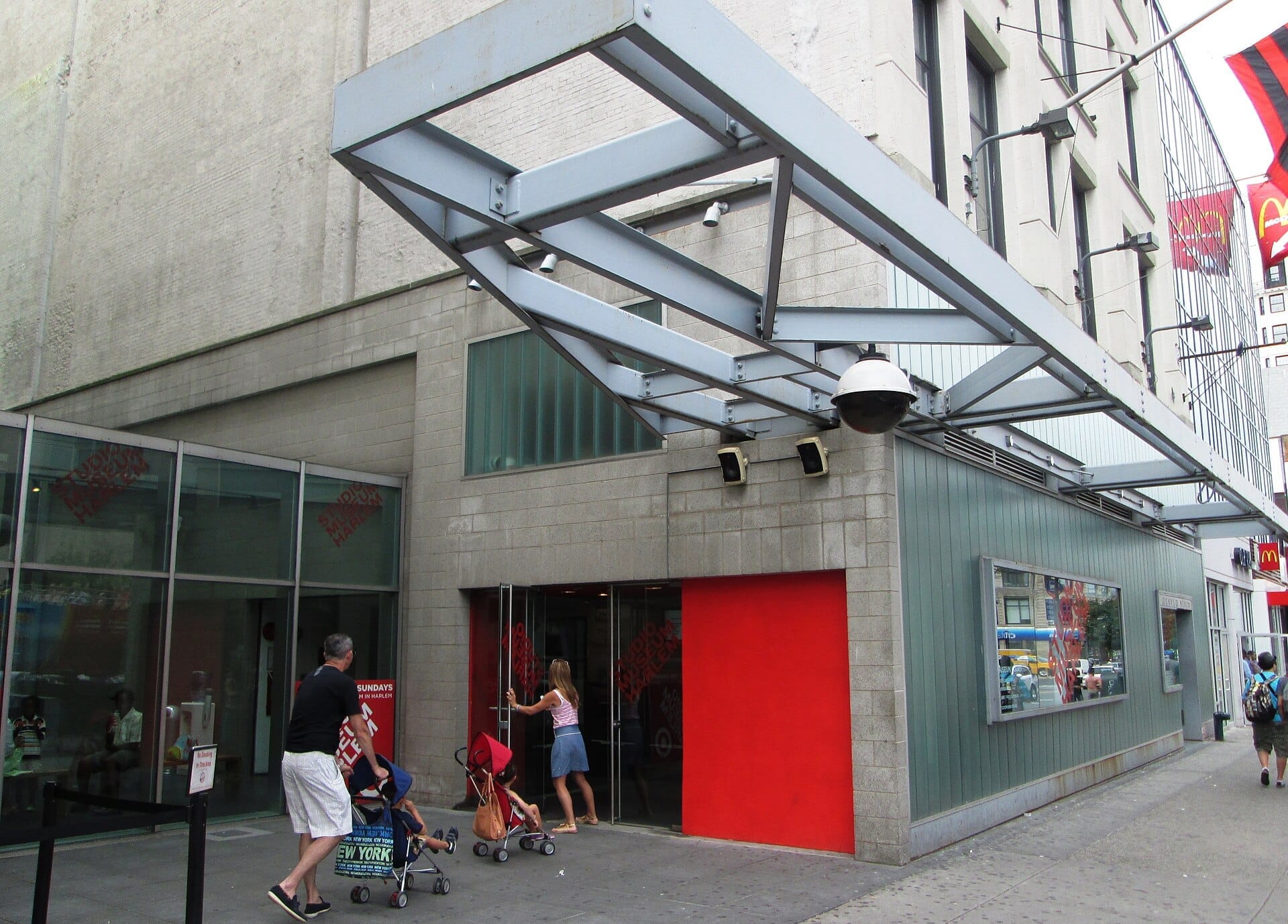 Vista esterna dello Studio Museum in Harlem, con architettura moderna, una vivace parete di accento rossa e persone che entrano su un marciapiede affollato.