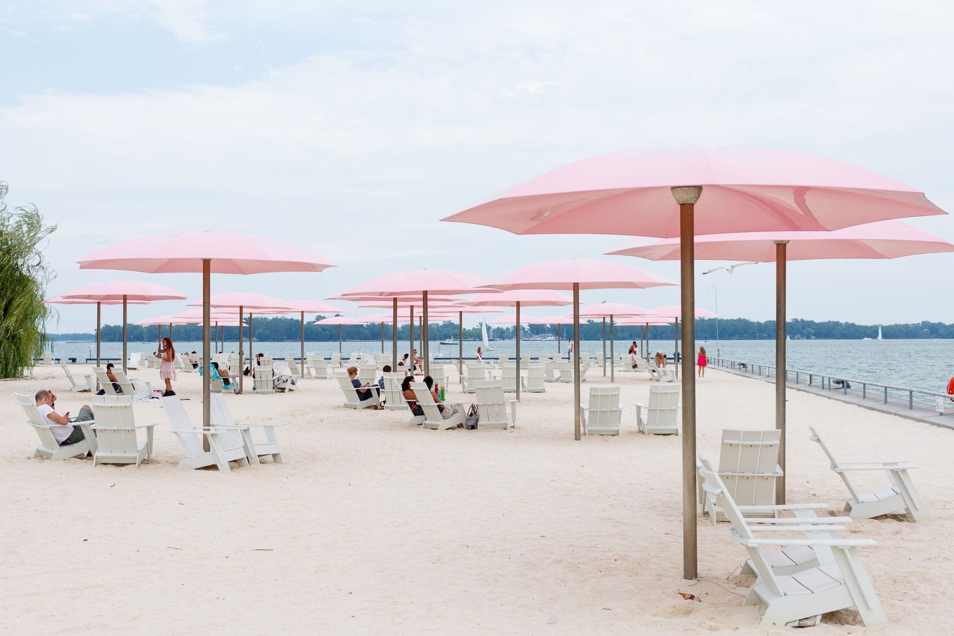 ชายหาดทรายในเมืองโตรอนโต พร้อมร่มชมพูไอคอนิคตั้งเรียงกัน เก้าอี้ขาว และผู้คนกำลังนั่งผ่อนคลายริมทะเลสาบ Ontario