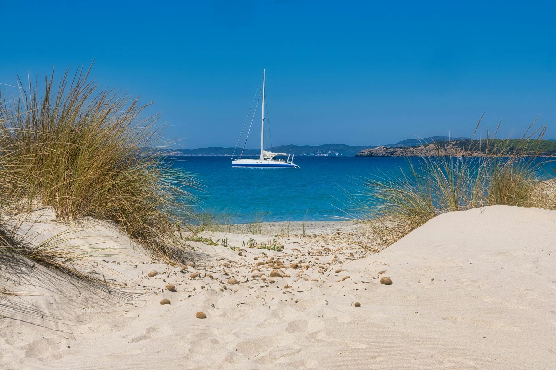 Weiße Sanddünen mit trockenem Gras in Porto Pino, Sulcis, mit Blick auf ein Katamaran auf dem klaren blauen Mittelmeer.