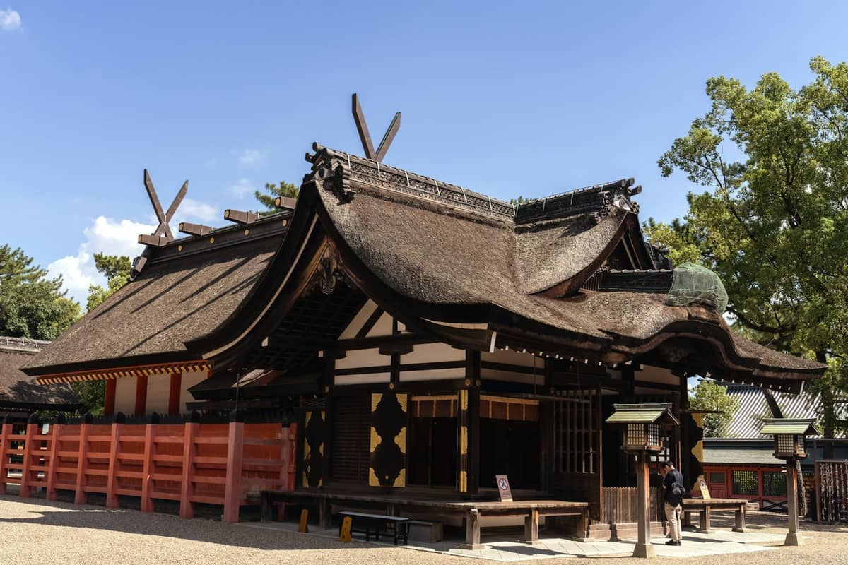 Sumiyoshi Taisha shrine in Osaka features traditional wooden architecture with a thatched roof, red fence, lanterns, and clear blue skies overhead.