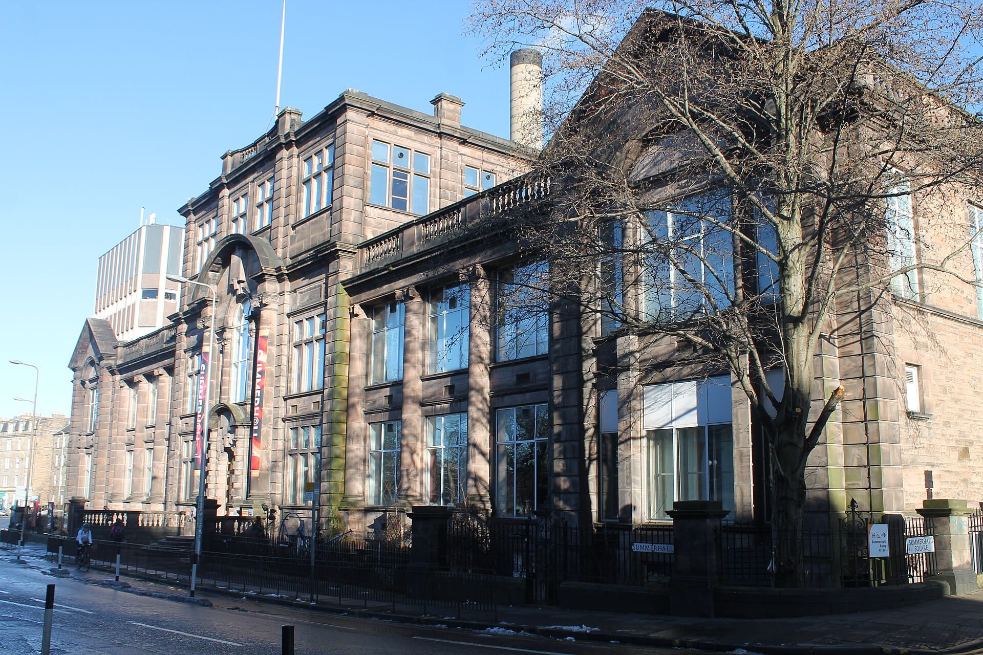Außenansicht von Summerhall in Edinburgh: die historische Gebäudefassade mit großen Fenstern, dem Eingangsbogen und Summerhall-Bannern an einem hellen, klaren Tag.