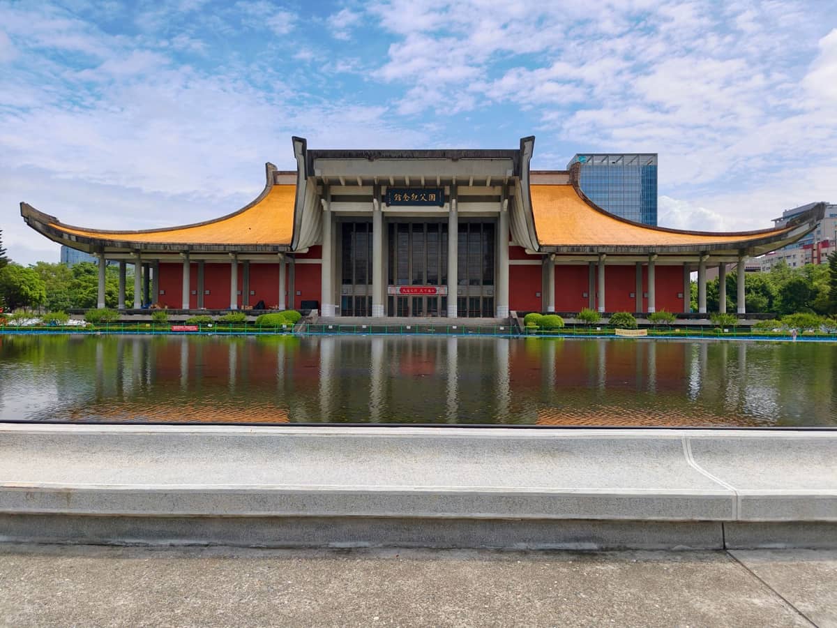 Weitwinkelansicht der National Dr. Sun Yat-sen Memorial Hall mit dem gelb gekachelten Dach und dem Spiegelteich davor unter blauem Himmel.