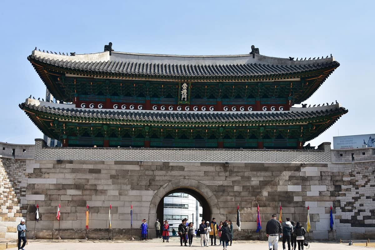 Vista frontal do Portão Sungnyemun (Namdaemun) em Seul, mostrando a base de pedra histórica, o elaborado pavilhão de madeira e visitantes na entrada sob um céu azul e limpo.