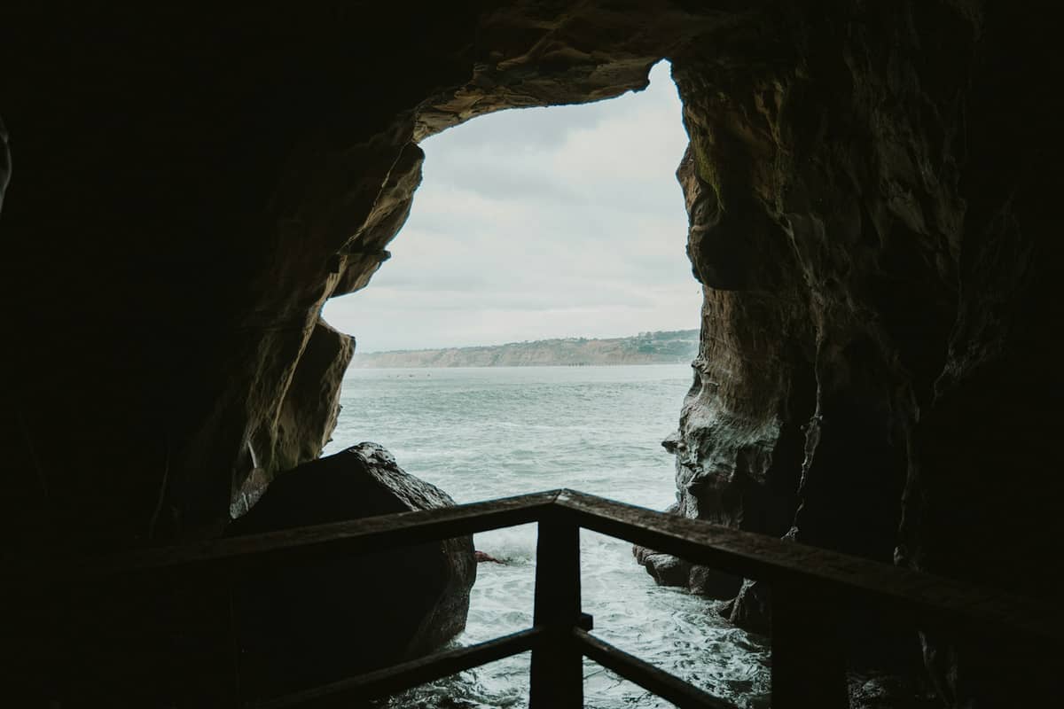 Blick aus dem Inneren der Sunny Jim Sea Cave nach draußen – durch die bogenförmige Öffnung sieht man den Ozean und die Küste in der Ferne, im Vordergrund ein Holzgeländer.