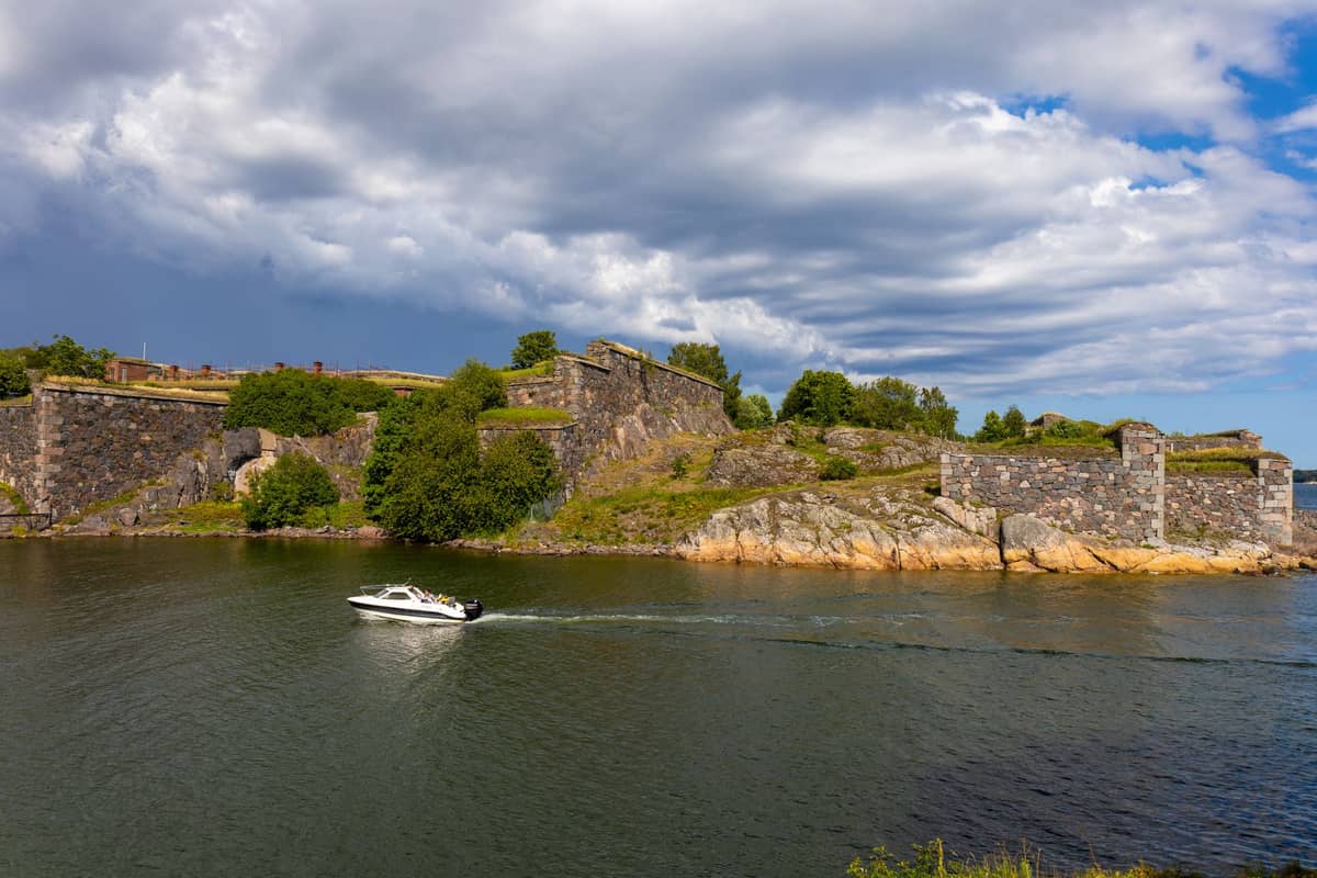 Muros de pedra da Fortaleza de Suomenlinna com árvores verdes e um barco passando pela água ao redor em um dia parcialmente nublado.