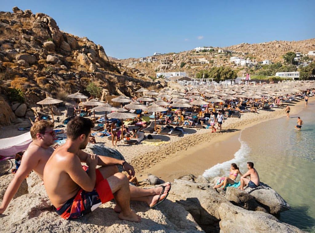 Vista de Super Paradise Beach en Mykonos con tumbonas, sombrillas de paja, personas descansando sobre las rocas y colinas doradas bajo un cielo azul despejado.