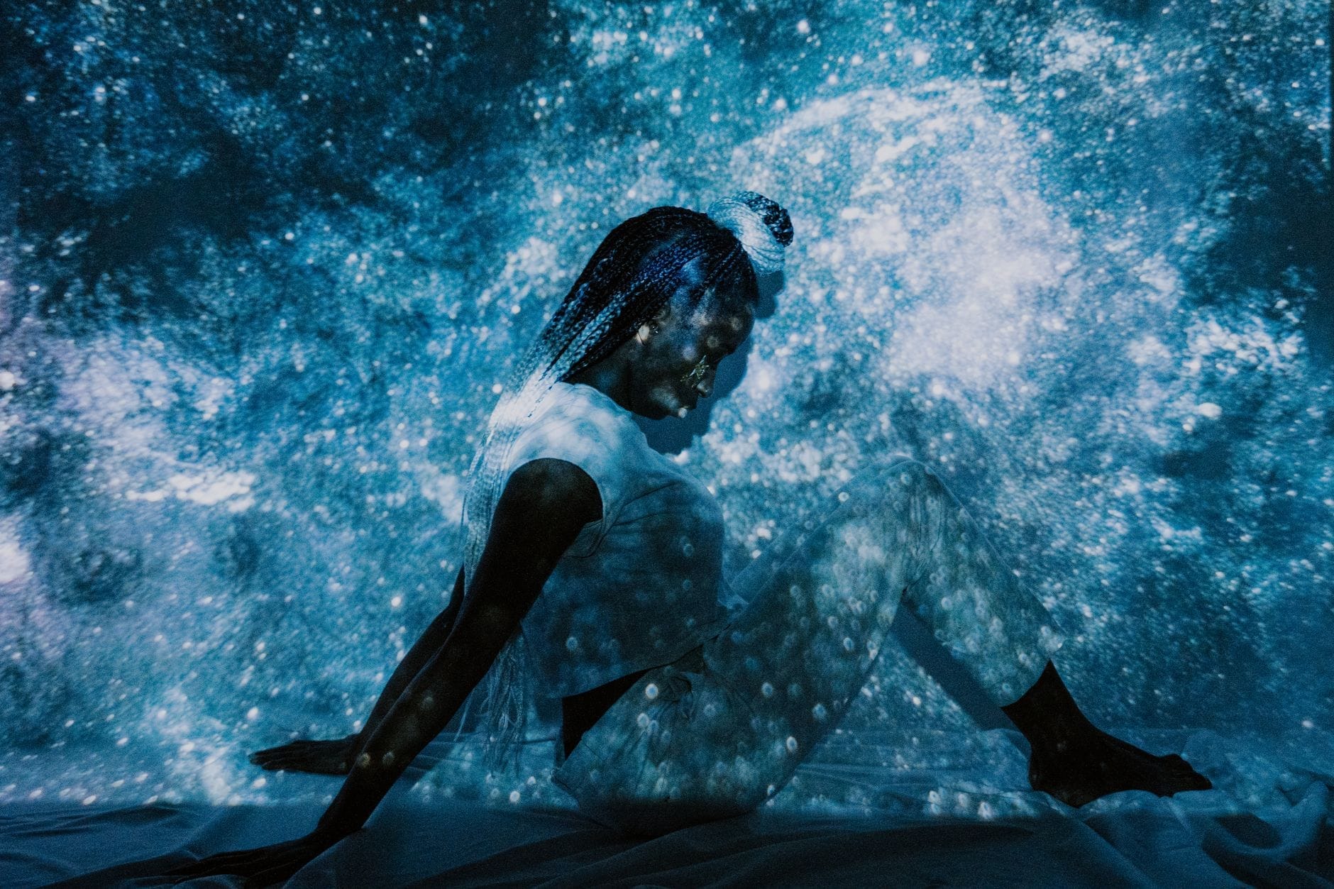 Una persona vestita di bianco seduta in una stanza buia, circondata da proiezioni di luce vorticosa blu e bianca che creano un'esperienza d'arte immersiva ipnotica.