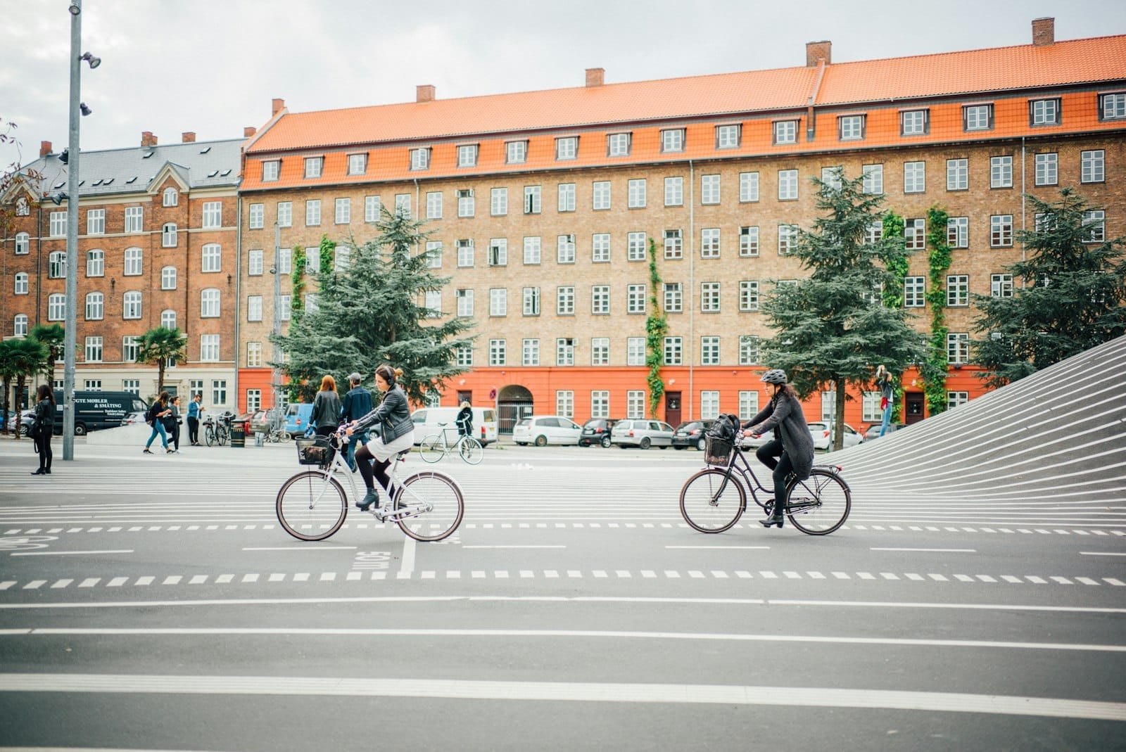 Ciclisti e pedoni si muovono nel parco Superkilen a Copenaghen, con pavimentazione a strisce, alberi sempreverdi ed edifici in mattoni sullo sfondo.