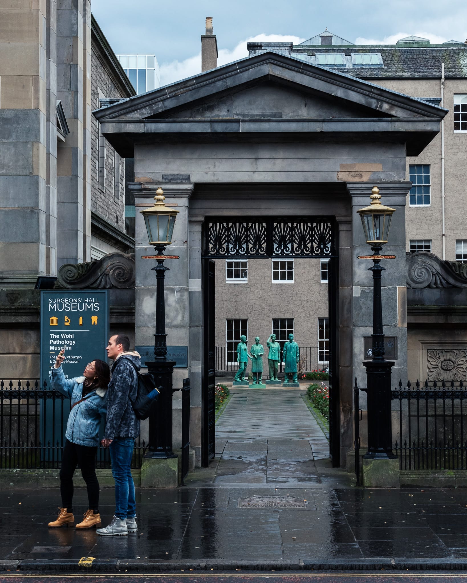 Eingang zu den Surgeons' Hall Museums in Edinburgh, mit einem neoklassizistischen Steintor, eisernen Laternen und grünen Statuen von Chirurgen im Innenhof.