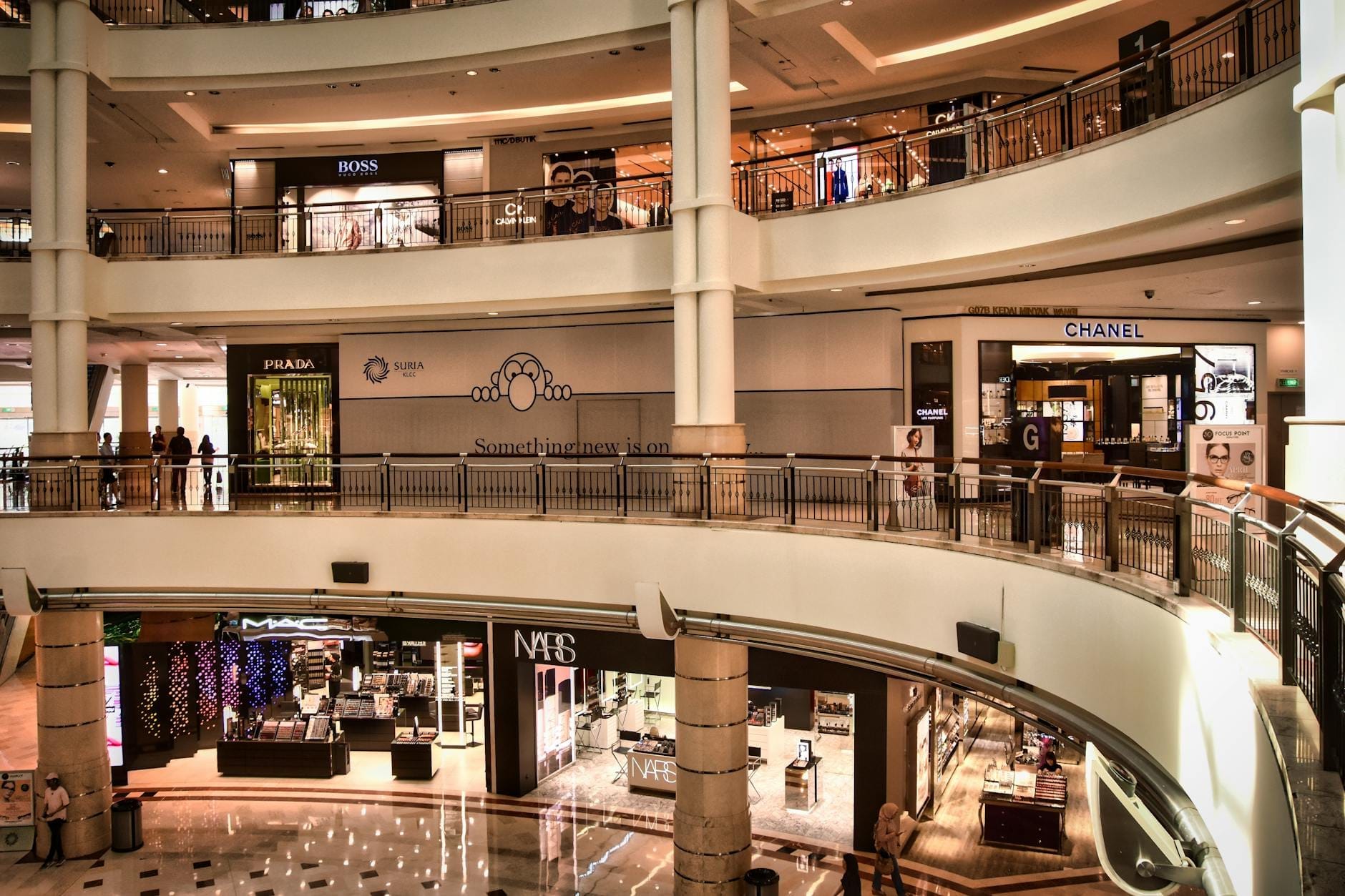 Interior view of Suria KLCC shopping mall with multiple luxury brand storefronts, curved walkways, and shoppers moving on different levels.