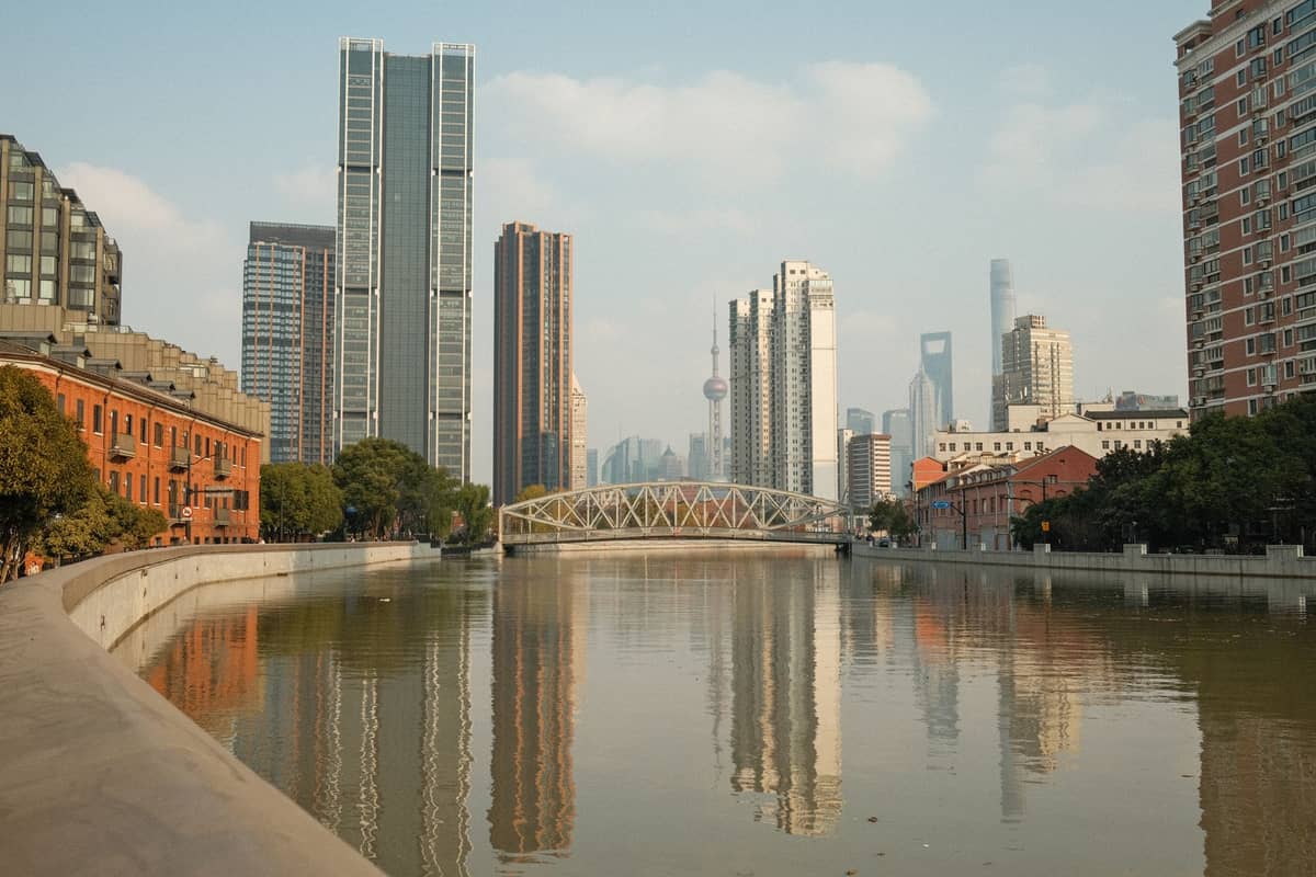 Weitläufige Ansicht der Uferpromenade am Suzhou Creek in Shanghai mit dem Creek, der Promenade, modernen Wolkenkratzern, historischen Backsteingebäuden und einer alten Eisenbrücke in der Ferne.