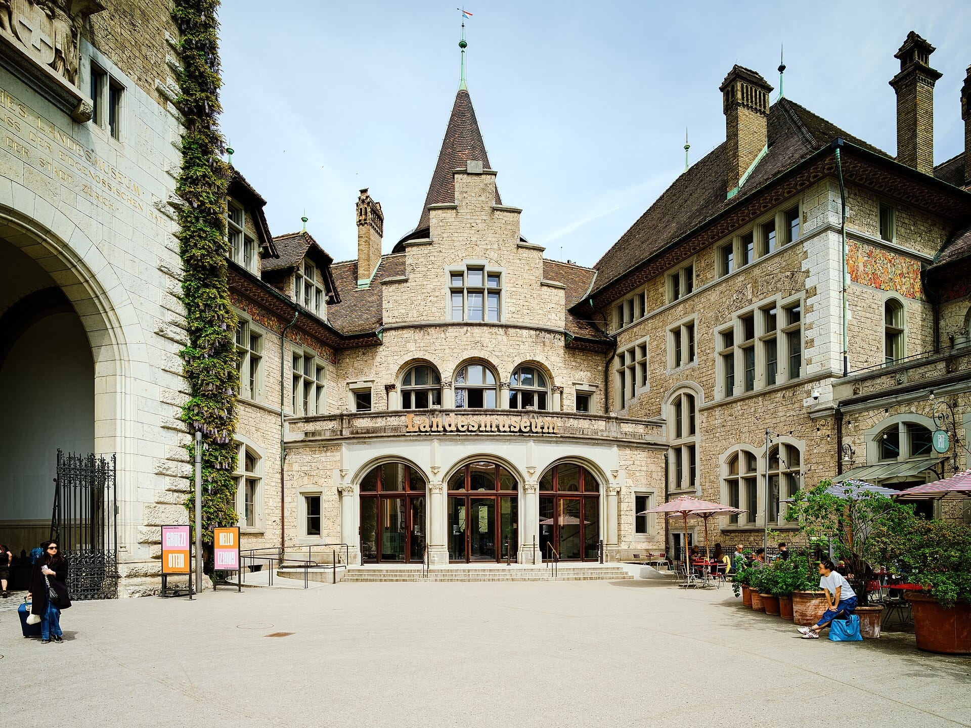 ด้านหน้าของ Swiss National Museum ในเมืองซูริก มีหน้าต่างโค้ง กำแพงหิน และผู้คนพักผ่อนในลานกลางแจ้งที่แดดจ้า