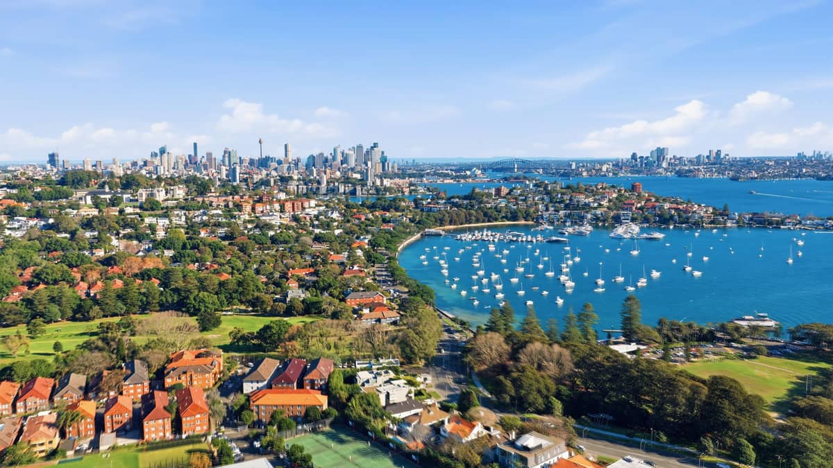 Luftaufnahme des Sydney Harbour mit grünen Landzungen, Buchten mit vertäuten Booten und der Skyline der Stadt in der Ferne unter blauem Himmel.