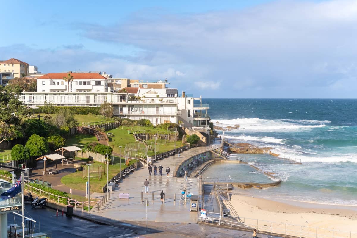 Nadmorska trasa spacerowa Bondi–Coogee w Sydney z ludźmi idącymi ścieżką wzdłuż oceanu i Bondi Icebergs w tle.