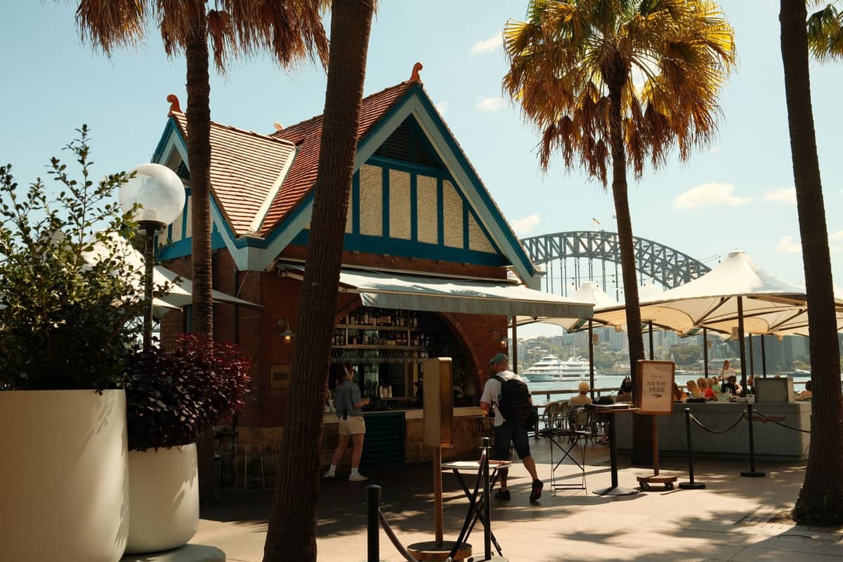 Café im Freien in Sydney mit Blick auf die Sydney Harbour Bridge, Palmen und Menschen, die an einem Kiosk Kaffee bestellen.