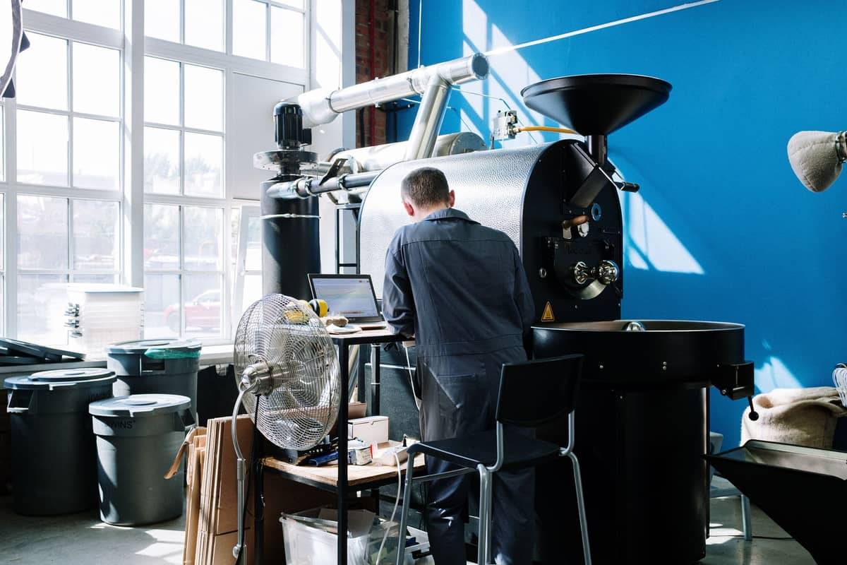 Person, die an einer großen Kaffeeröstmaschine in einer hellen, modernen Rösterei mit blauen Wänden und großen Fenstern arbeitet.