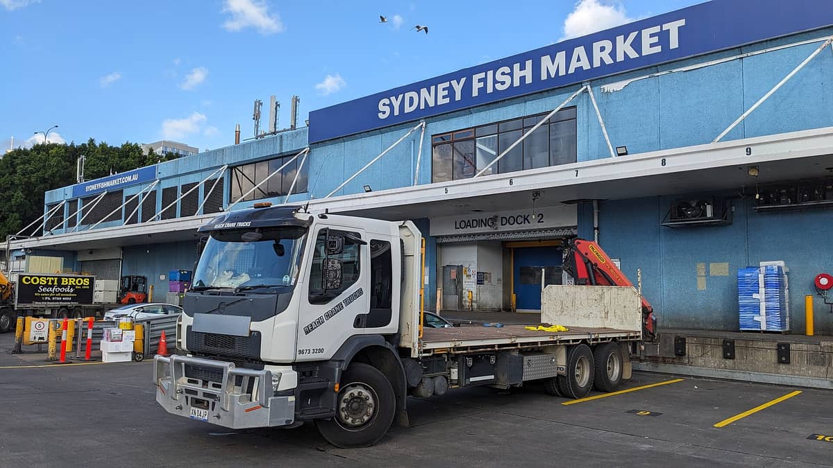 Vista amplia del exterior del Mercado de Pescado de Sídney con su gran fachada azul, el llamativo cartel con el nombre del mercado y un camión de reparto estacionado en el muelle de carga.