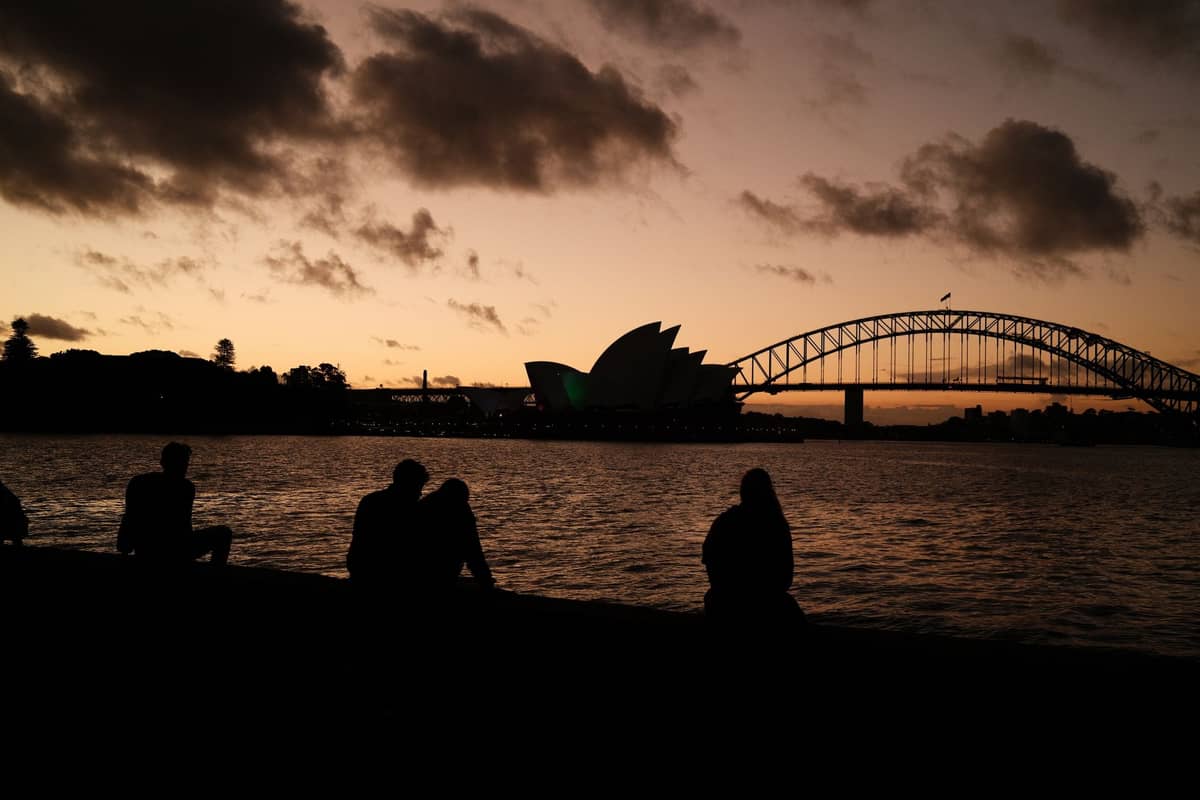 Silhouettierte Paare sitzen bei Sonnenuntergang am Sydney Harbour mit dem Sydney Opera House und der Harbour Bridge im Blick.